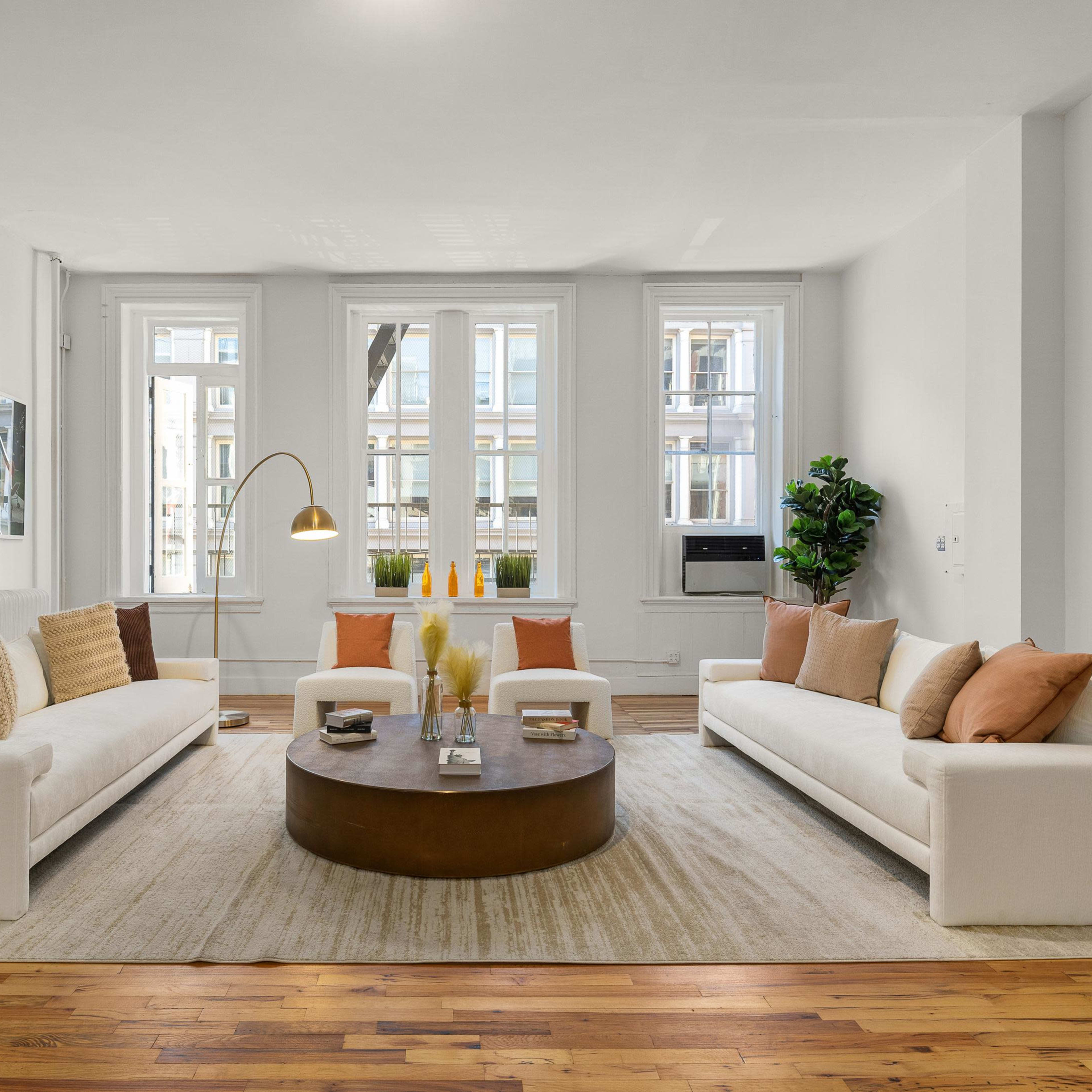 A bright living room with two white sofas facing a round coffee table, large windows, and a light-colored rug on a wooden floor.