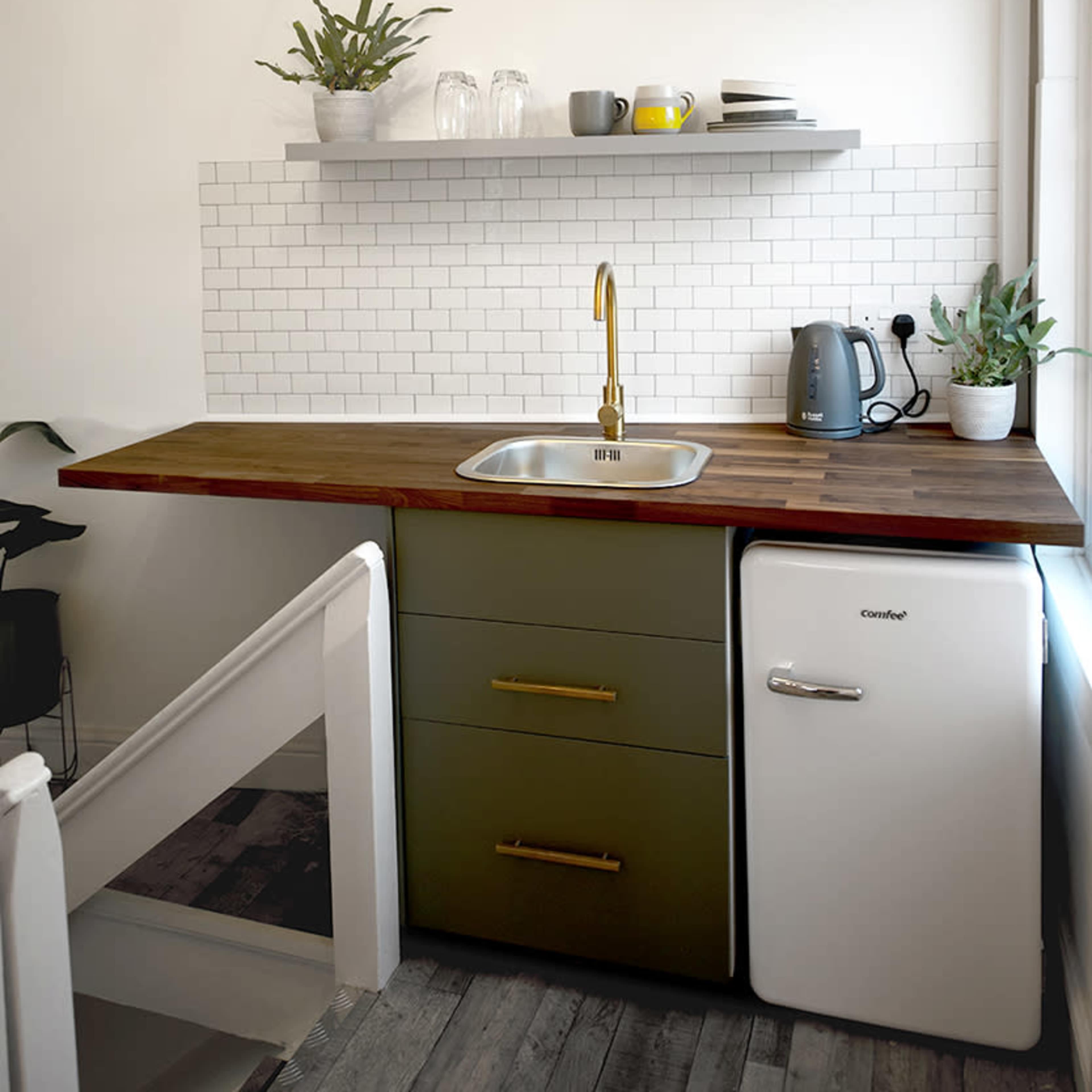 A compact kitchen featuring a wooden countertop, a white refrigerator, a stainless steel sink with a gold faucet, and a shelf with kitchenware and plants.
