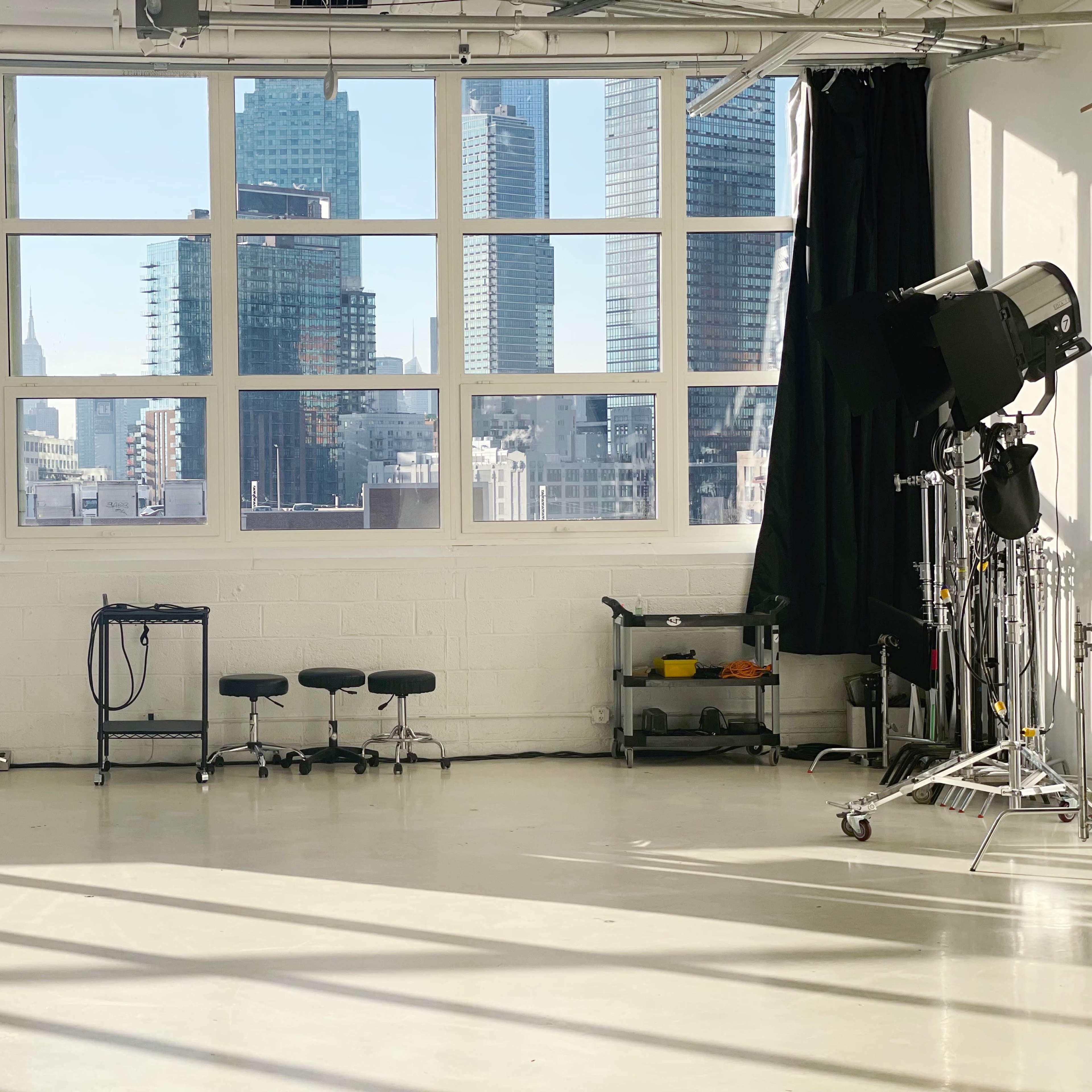 A well-lit studio space with large windows displaying a city skyline, black curtains, and equipment on wheels.