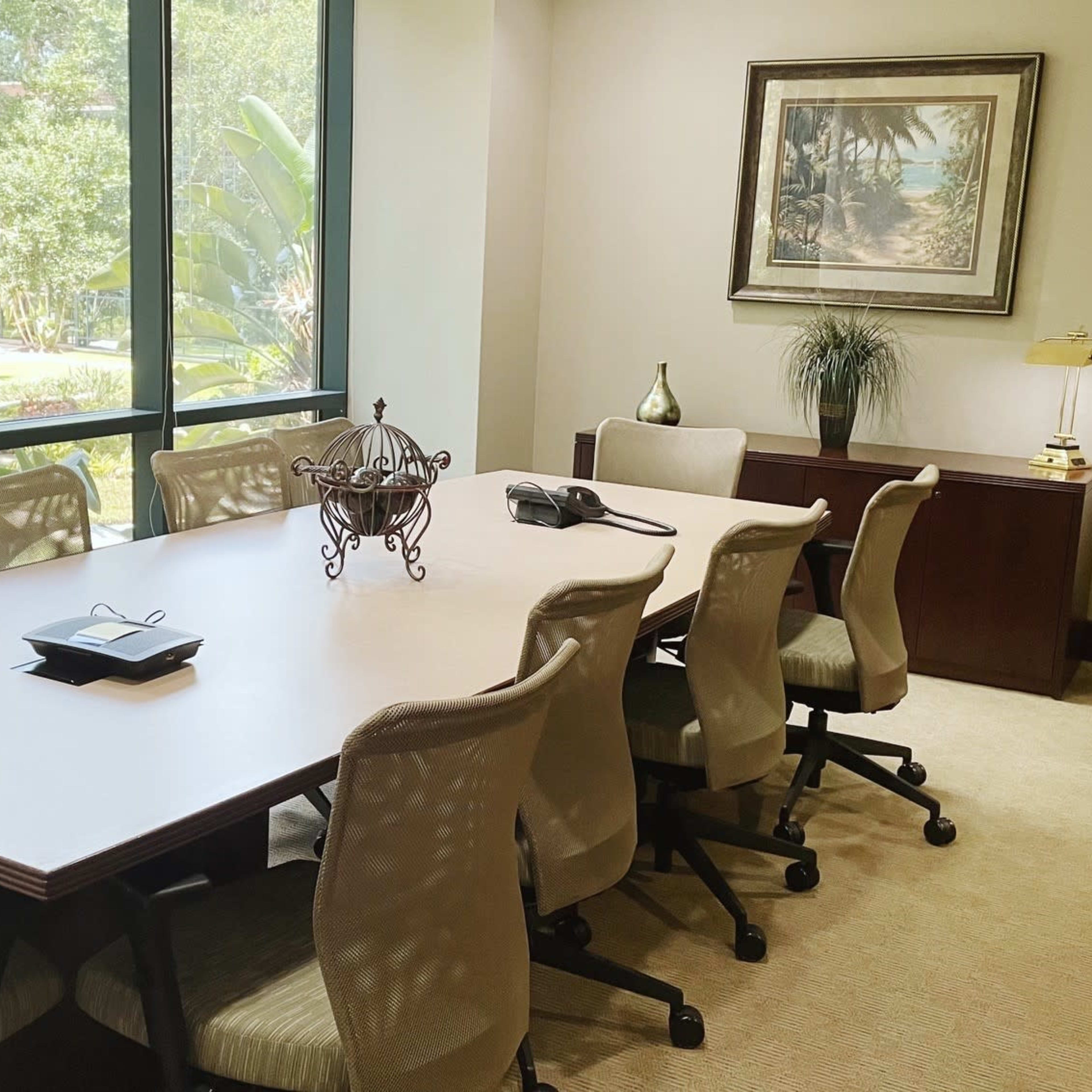 A conference room features a large table surrounded by several chairs, with a phone and decorative items on the table, and large windows overlooking greenery.