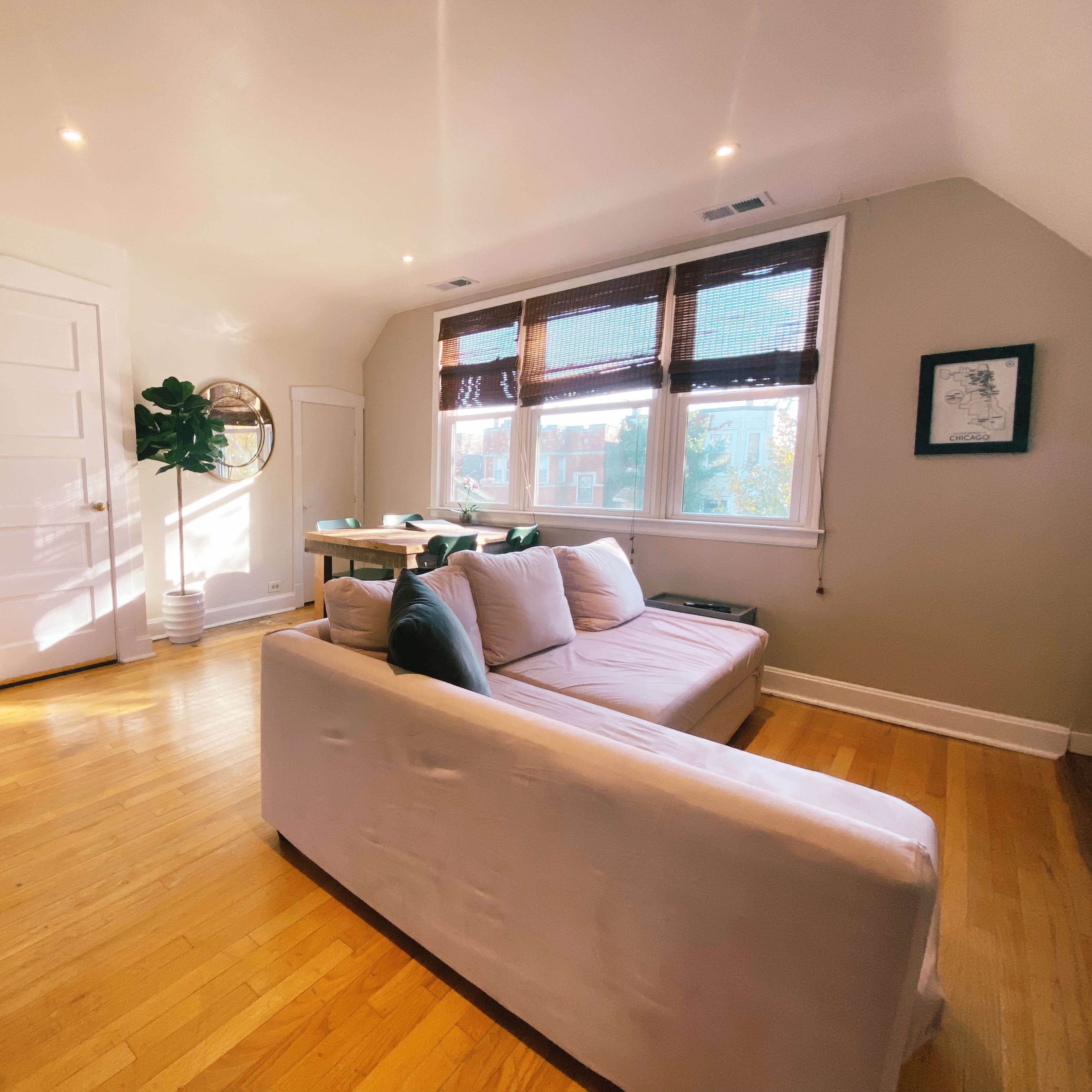 A cozy living room with a white couch, wooden flooring, and large windows covered by bamboo shades.