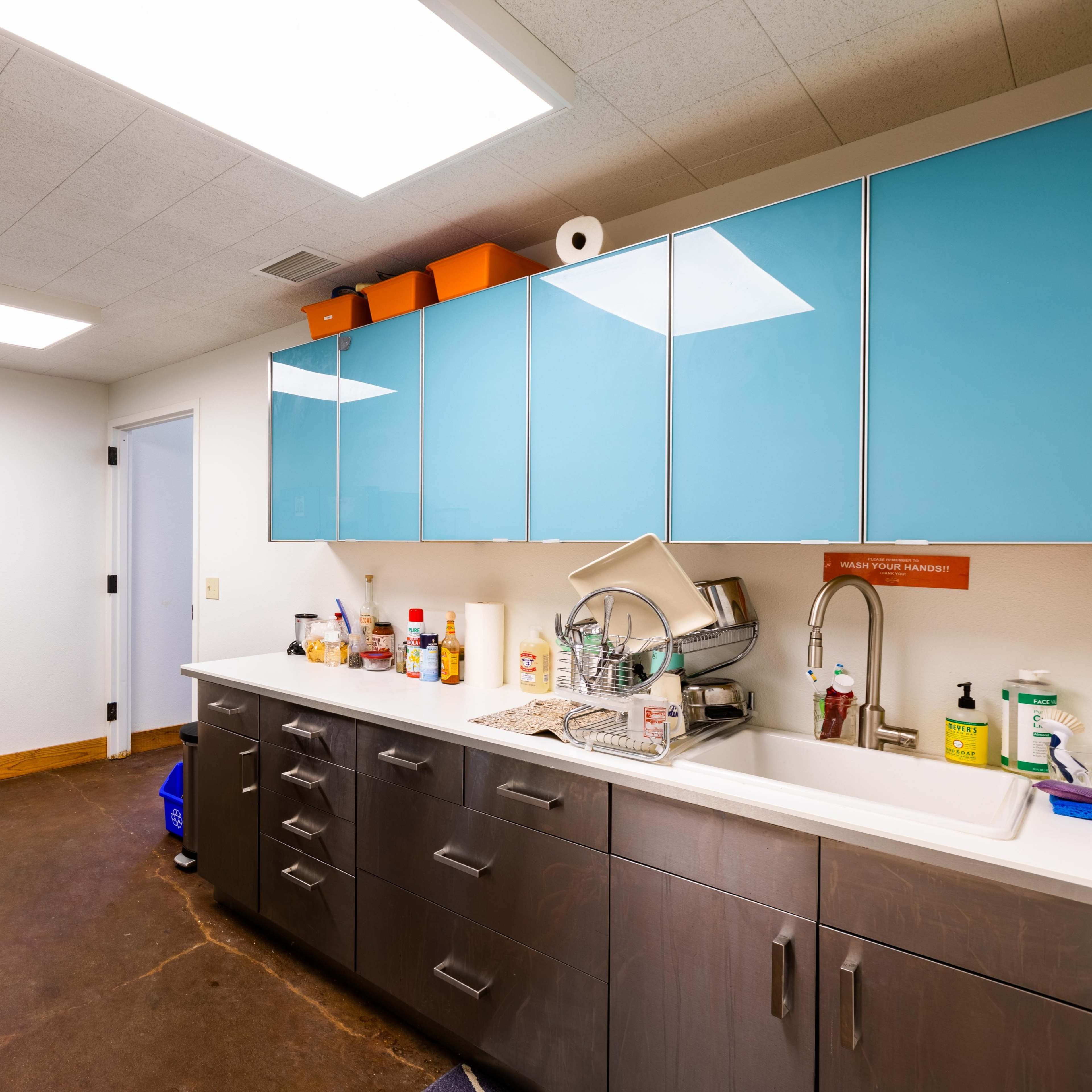 A kitchen with blue cabinets, a stainless steel sink, and appliances, featuring a concrete floor and overhead lighting.
