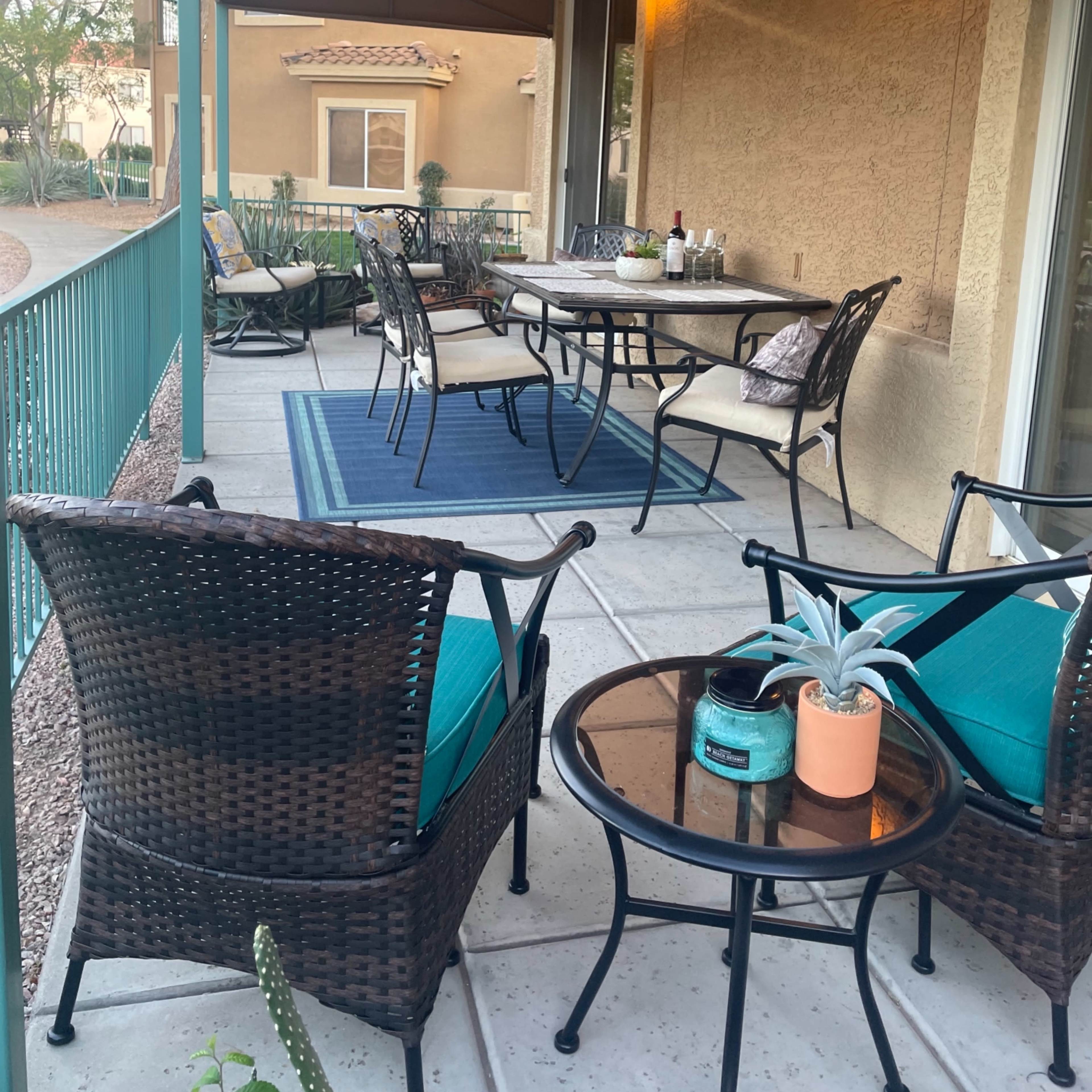 A covered outdoor patio with a dining table, several chairs, and a seating area featuring wicker furniture and a small potted plant.