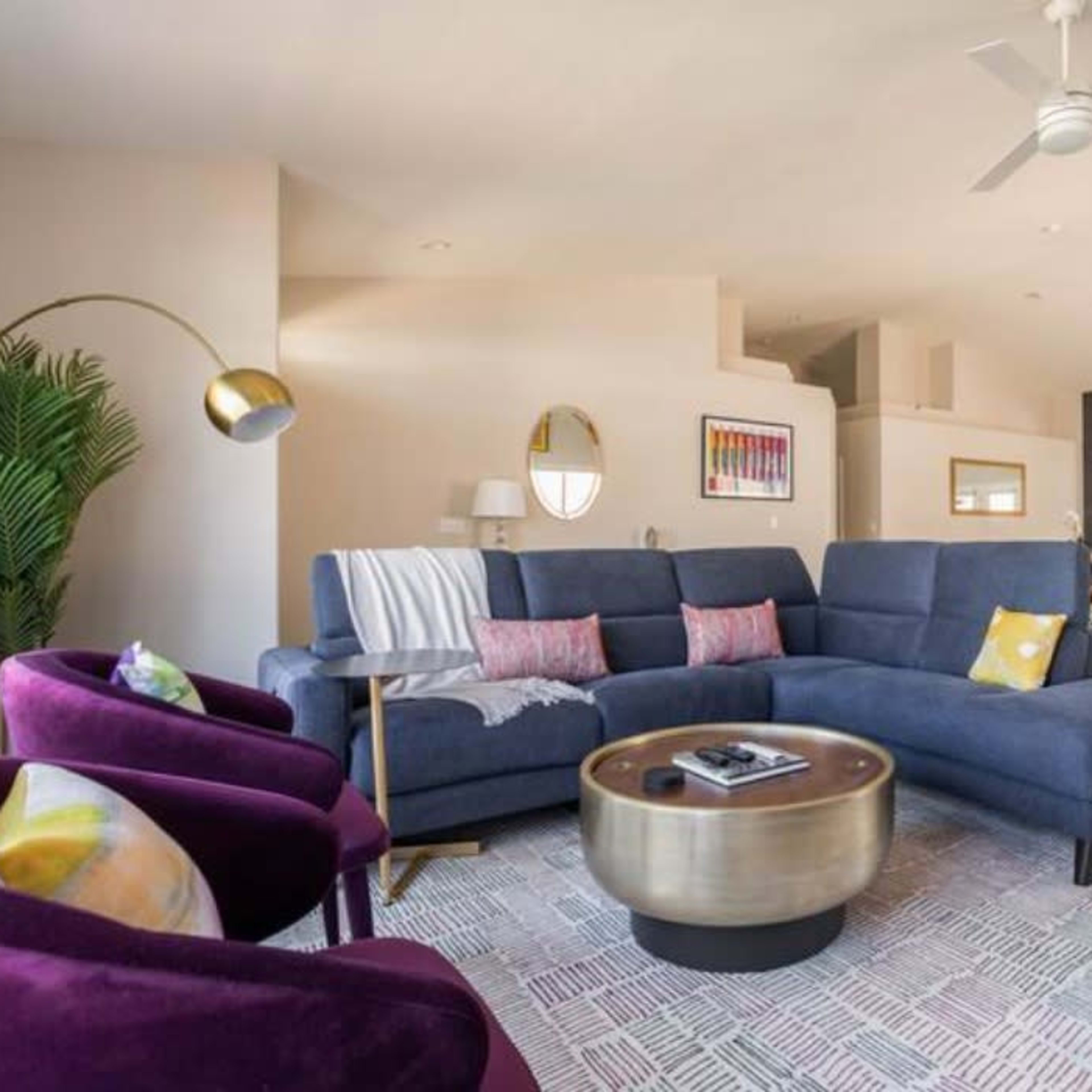 A modern living room features a blue sectional sofa, two purple armchairs, a round coffee table, and a patterned area rug, with large windows allowing natural light.