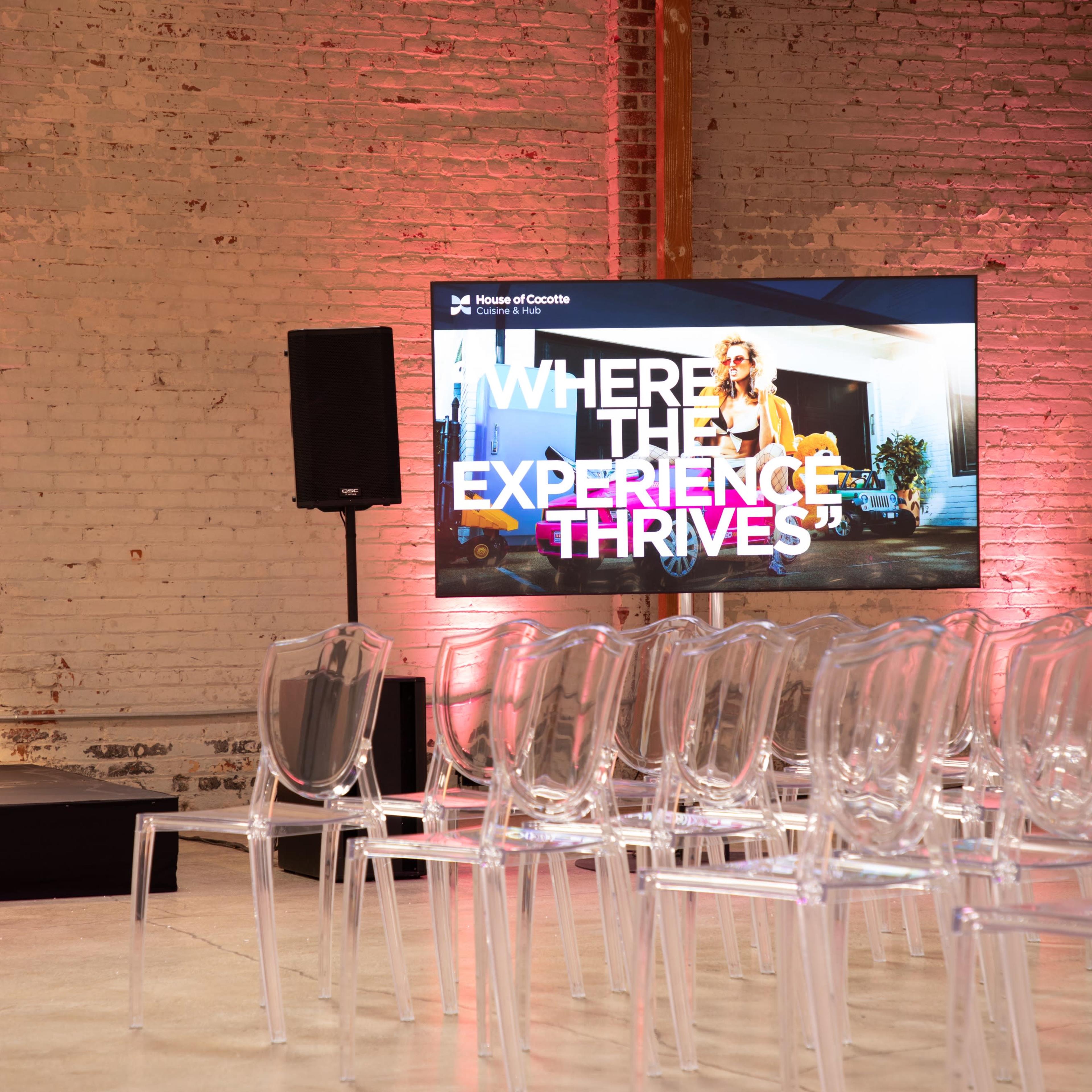 A spacious venue set up for an event, featuring transparent chairs arranged in front of a large screen displaying promotional content, with a illuminated podium to the side.