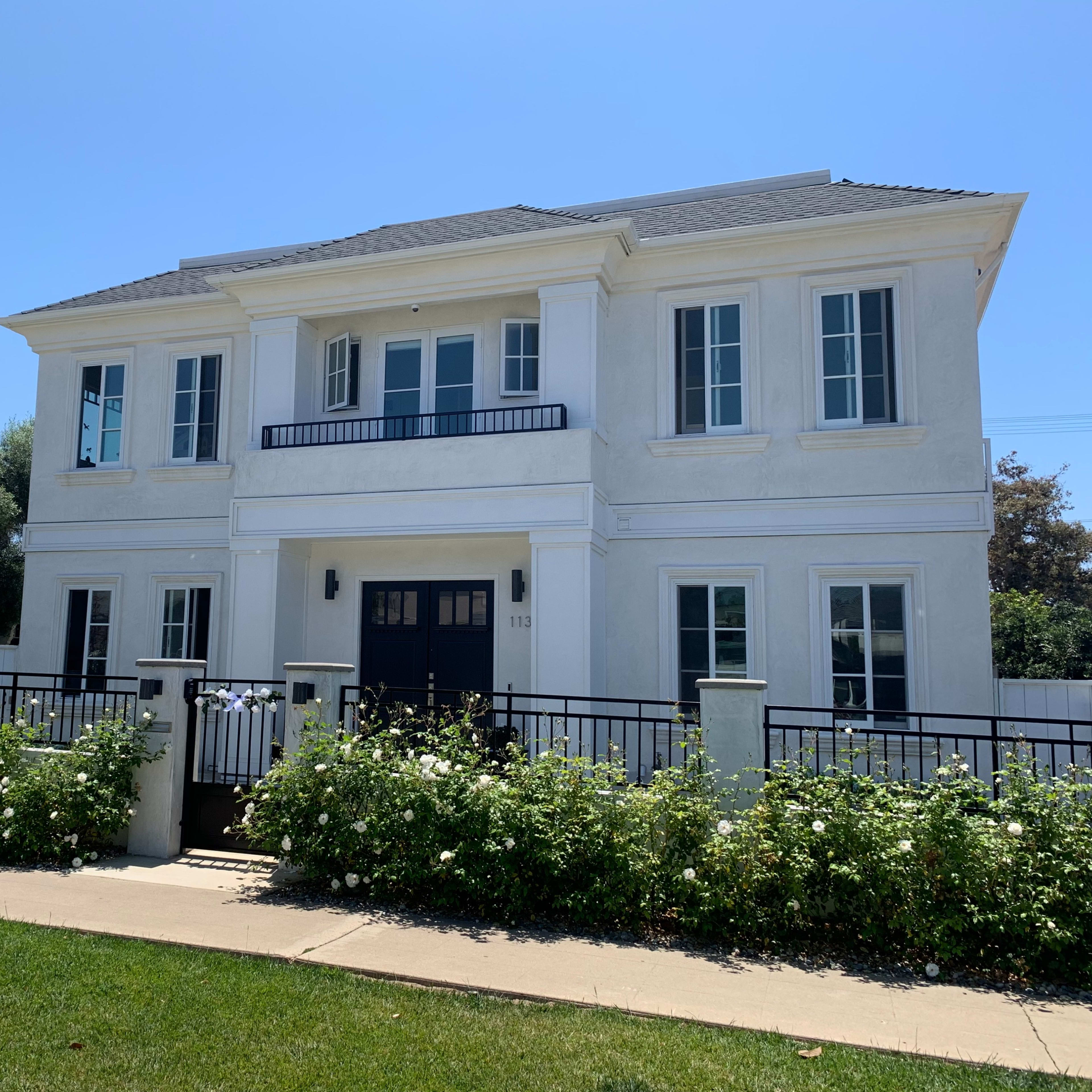 A two-story white house with black accents and a front balcony is surrounded by a well-maintained garden and lawn.