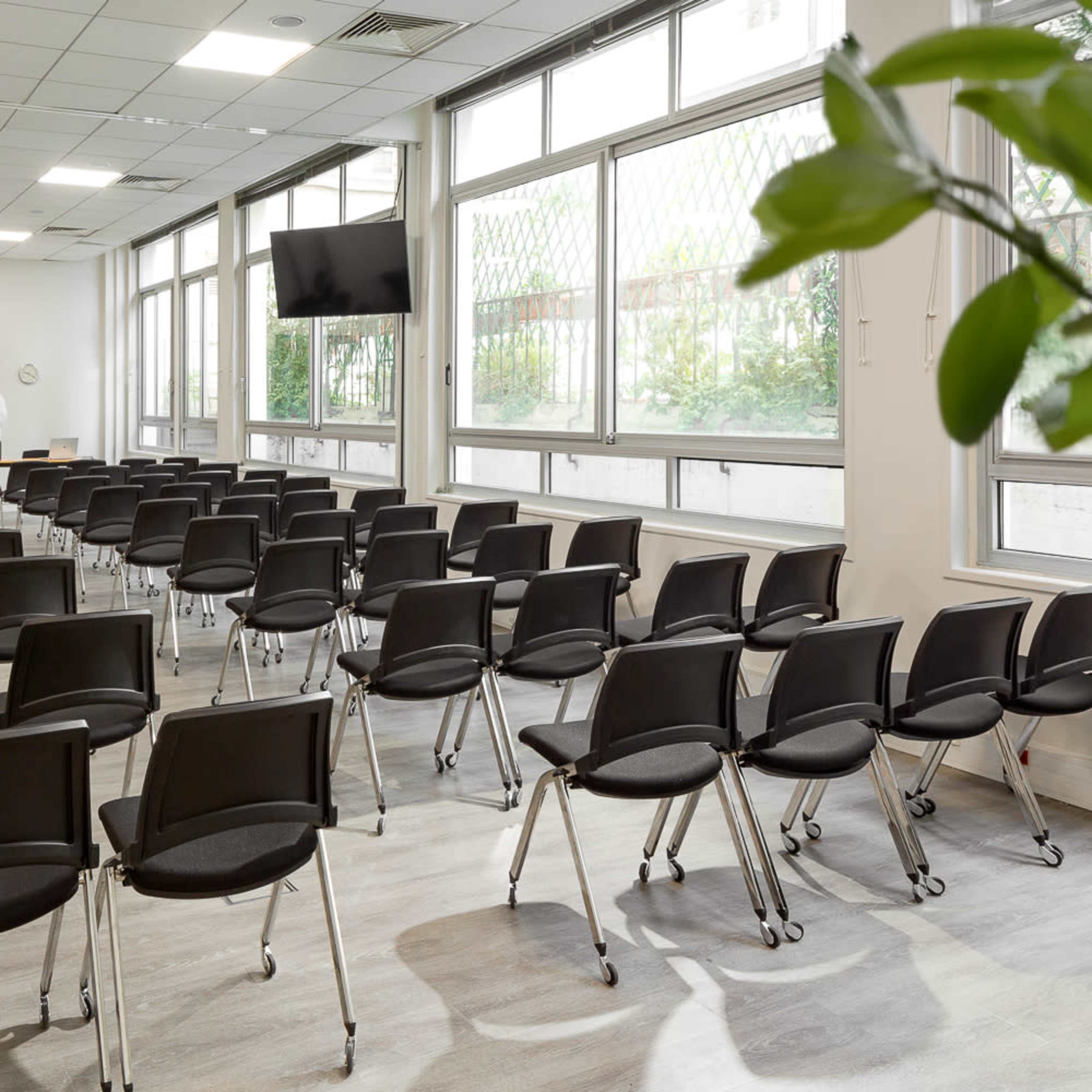 A modern conference room is set up with rows of empty black chairs facing a blank screen and two people conversing near the front.