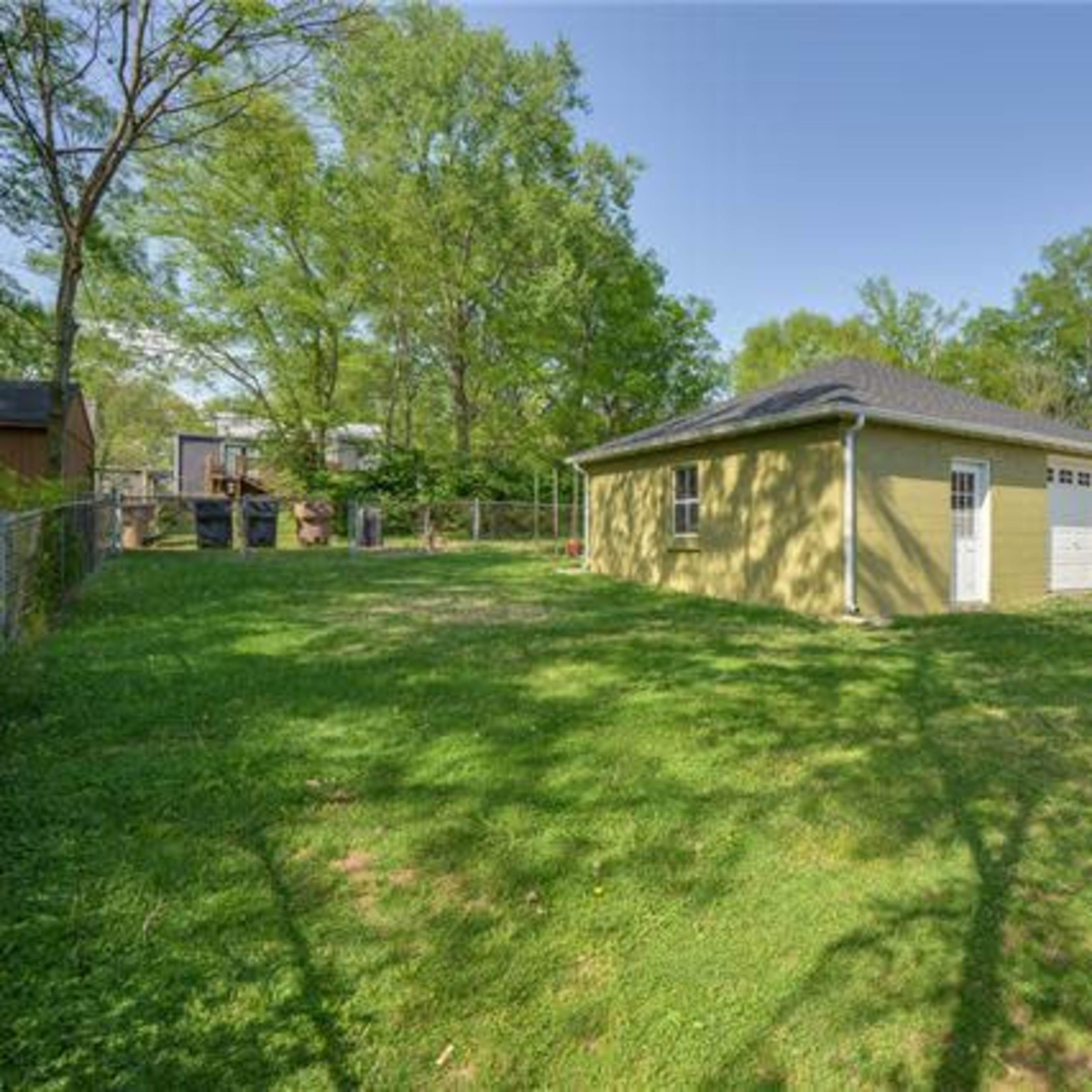 A yellow building with a garage door sits in a grassy backyard surrounded by trees and a wooden fence.