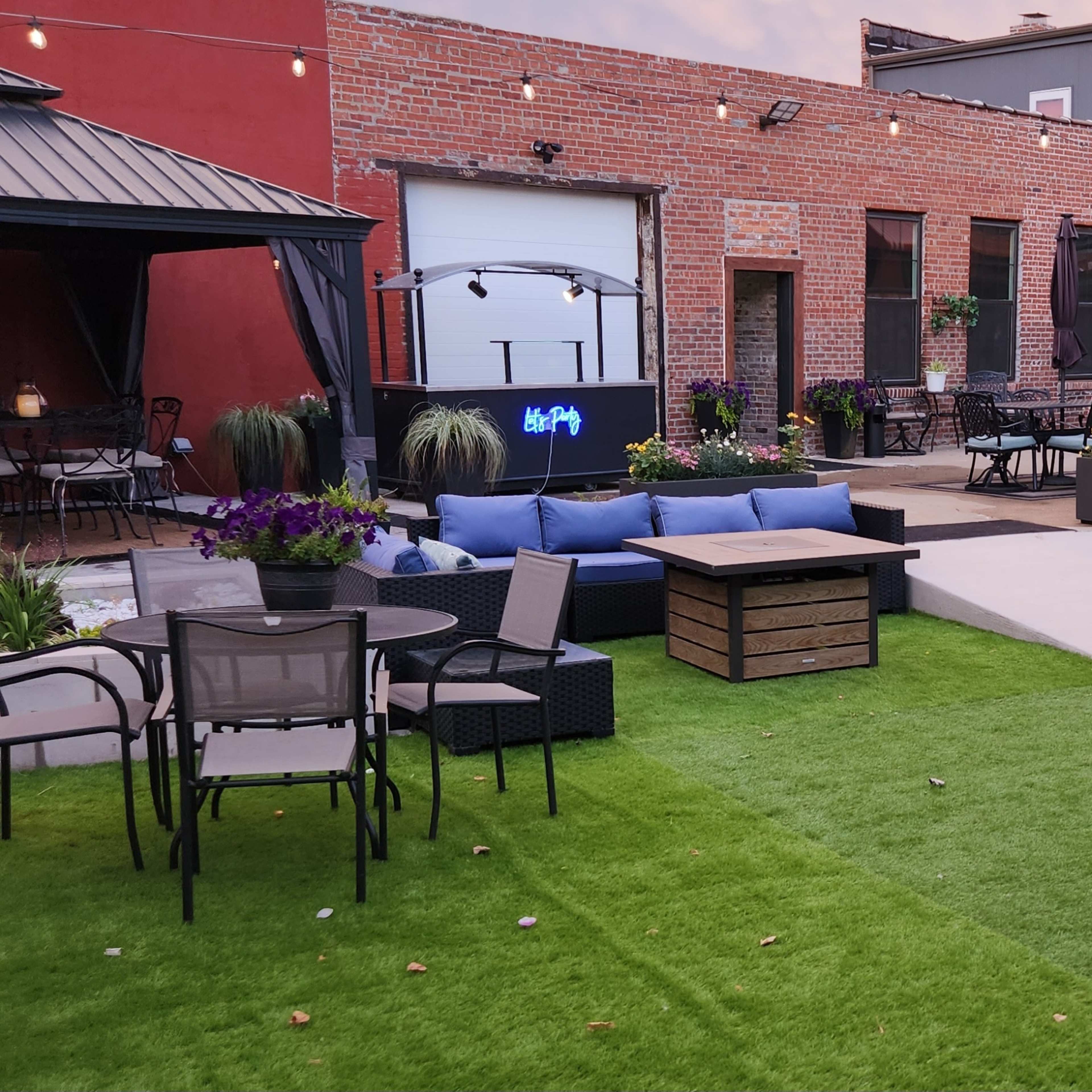 A landscaped outdoor patio area featuring artificial grass, a seating arrangement with sofas and chairs, potted plants, and string lights overhead.