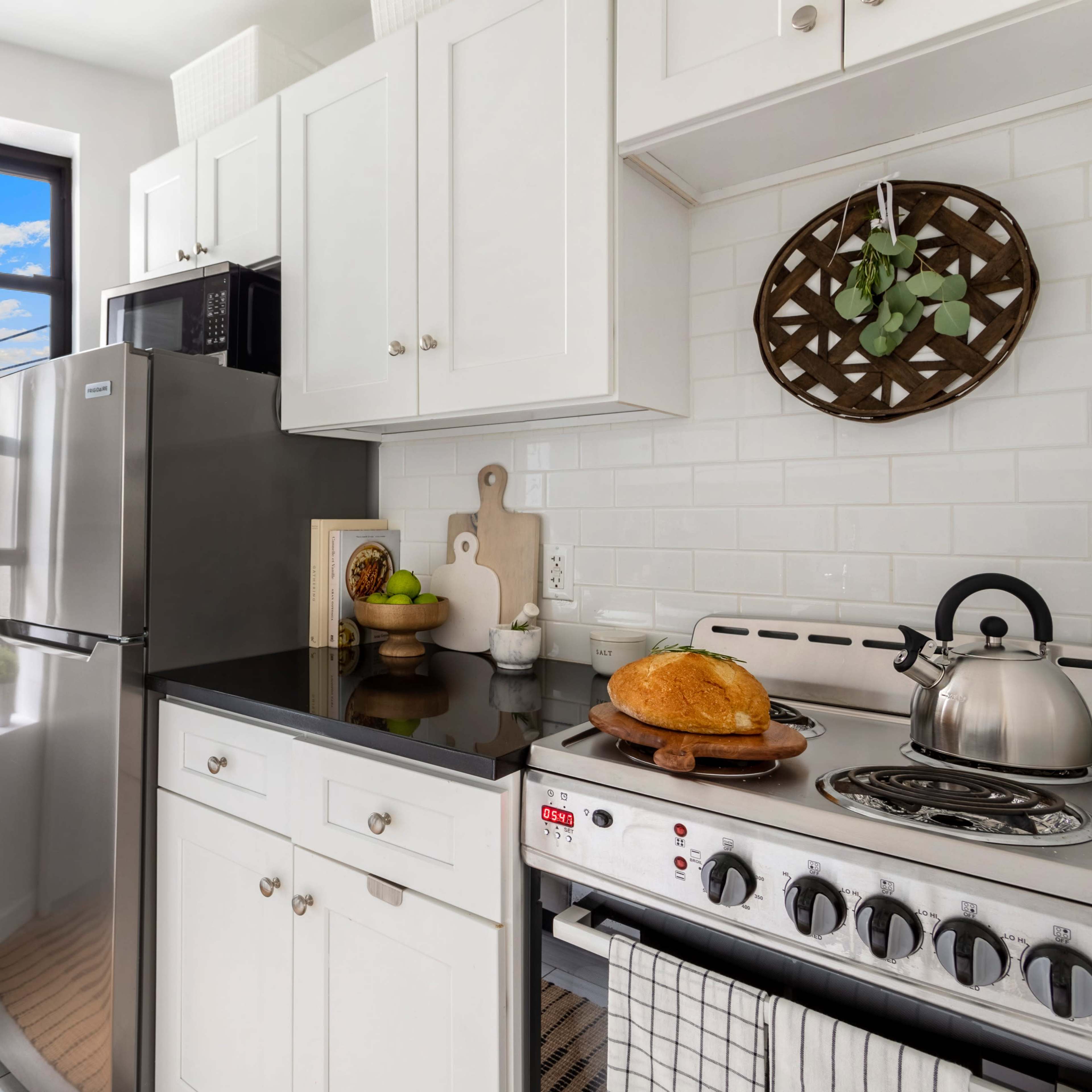 A compact kitchen features white cabinetry, a stainless steel refrigerator, and a stove with a loaf of bread on the counter beside a kettle.