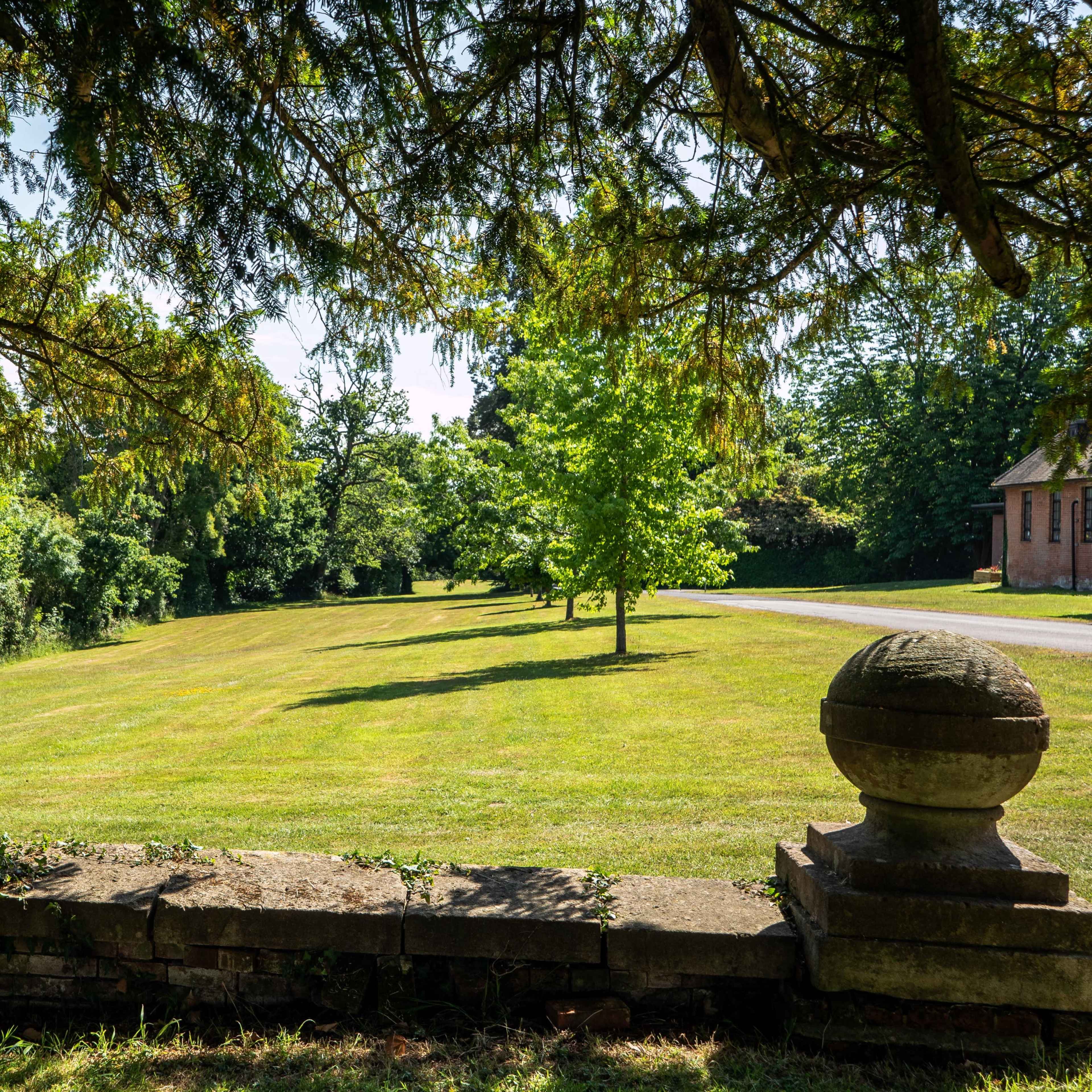 A grassy open area bordered by trees, with a stone wall in the foreground and a building visible in the background.