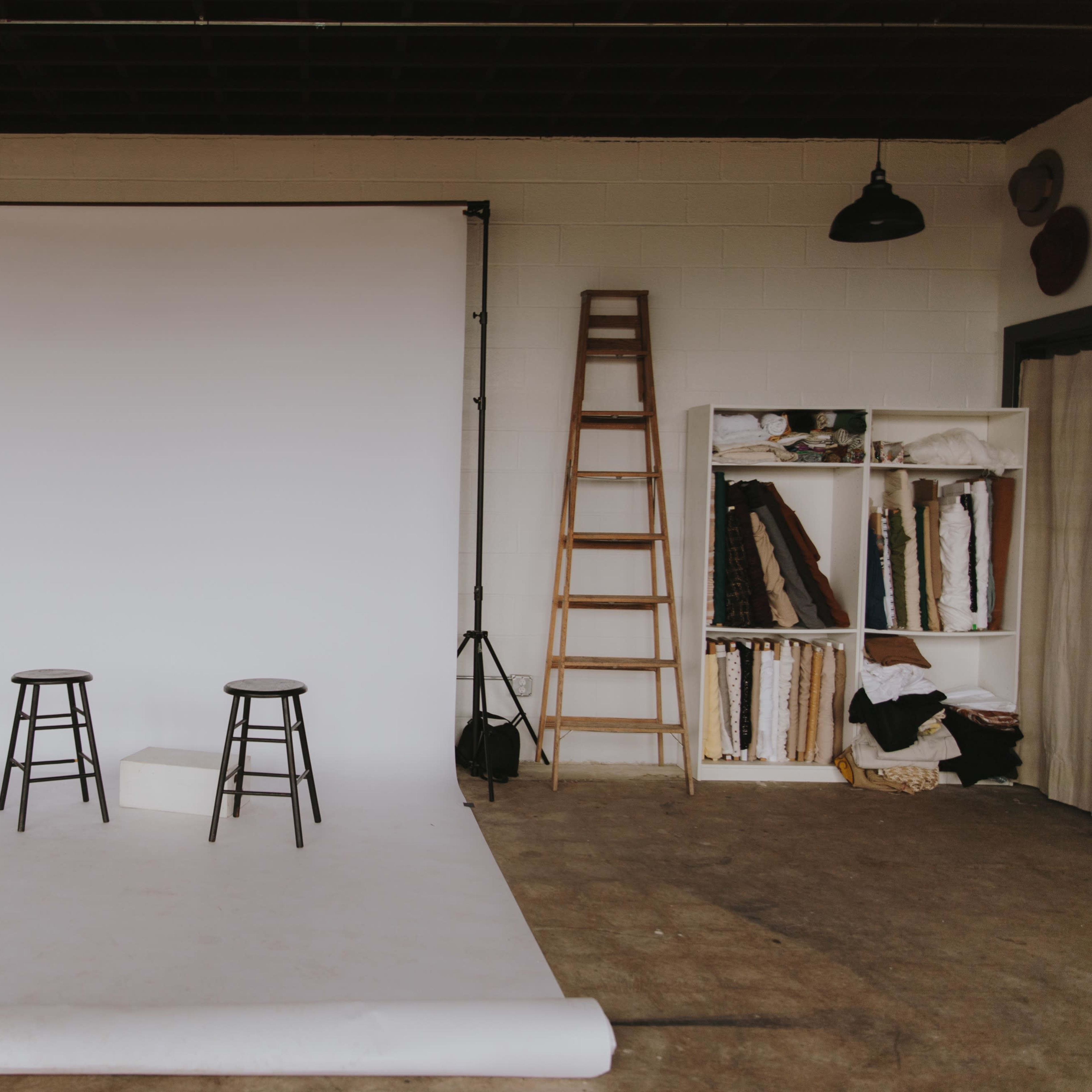 A photography studio with a white backdrop, two black stools, a ladder, and a shelving unit filled with fabric rolls.