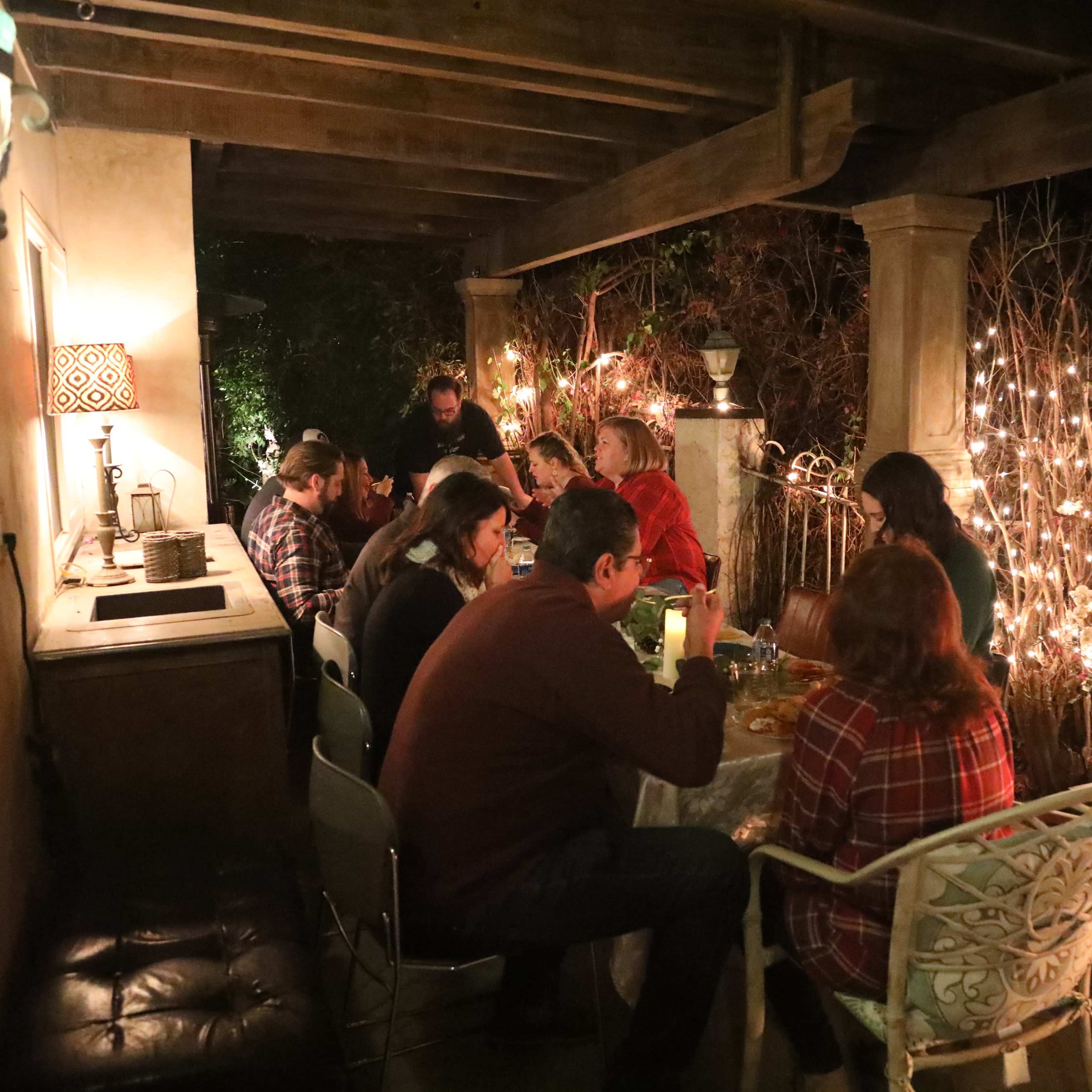 A group of people is seated around a table on a patio decorated with string lights and surrounded by a garden.