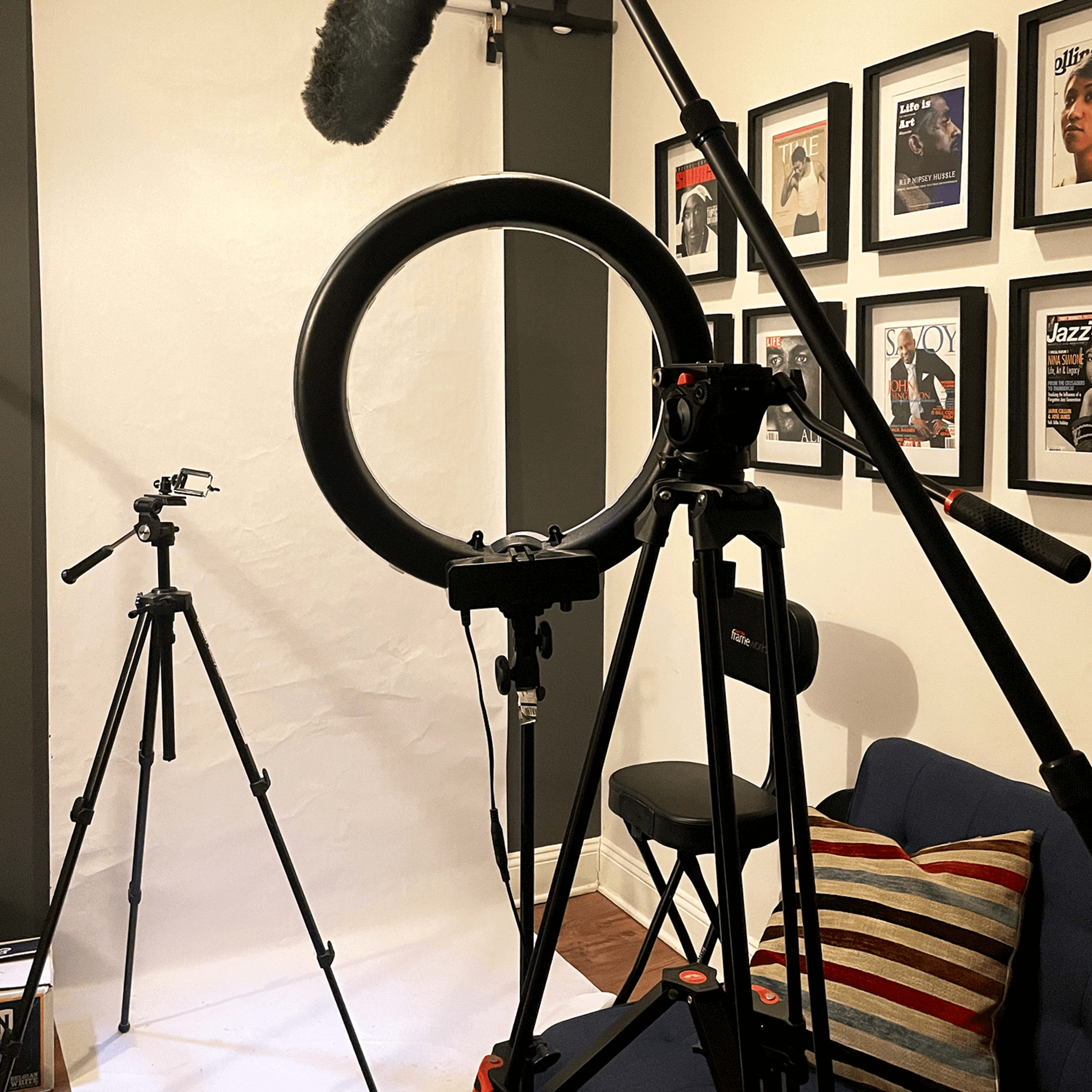 A studio setup with tripods, a microphone on a boom, a ring light, and a white backdrop in a room adorned with framed posters.