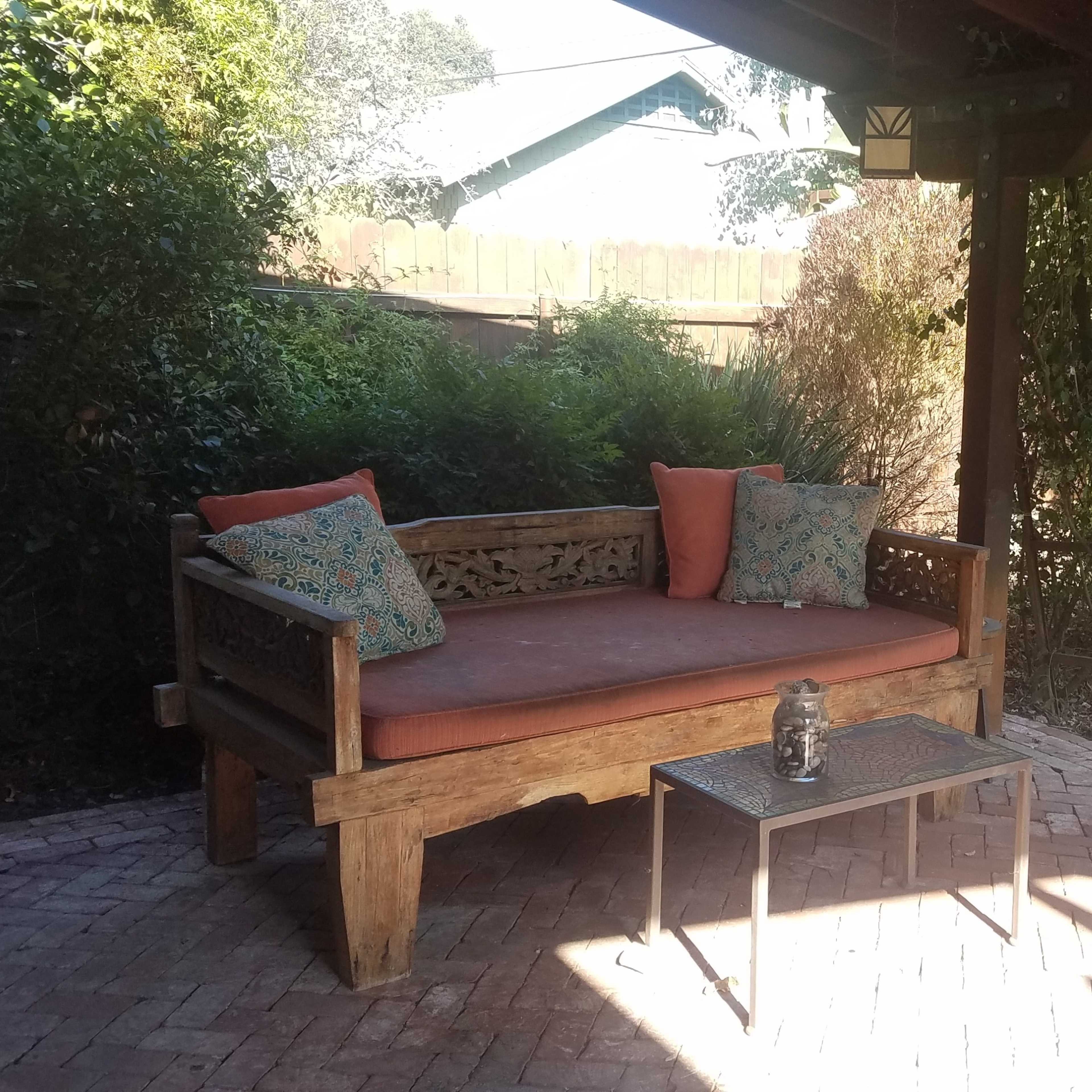 A shaded outdoor seating area with a wooden couch adorned with orange cushions, a small table, and an Adirondack chair surrounded by greenery.