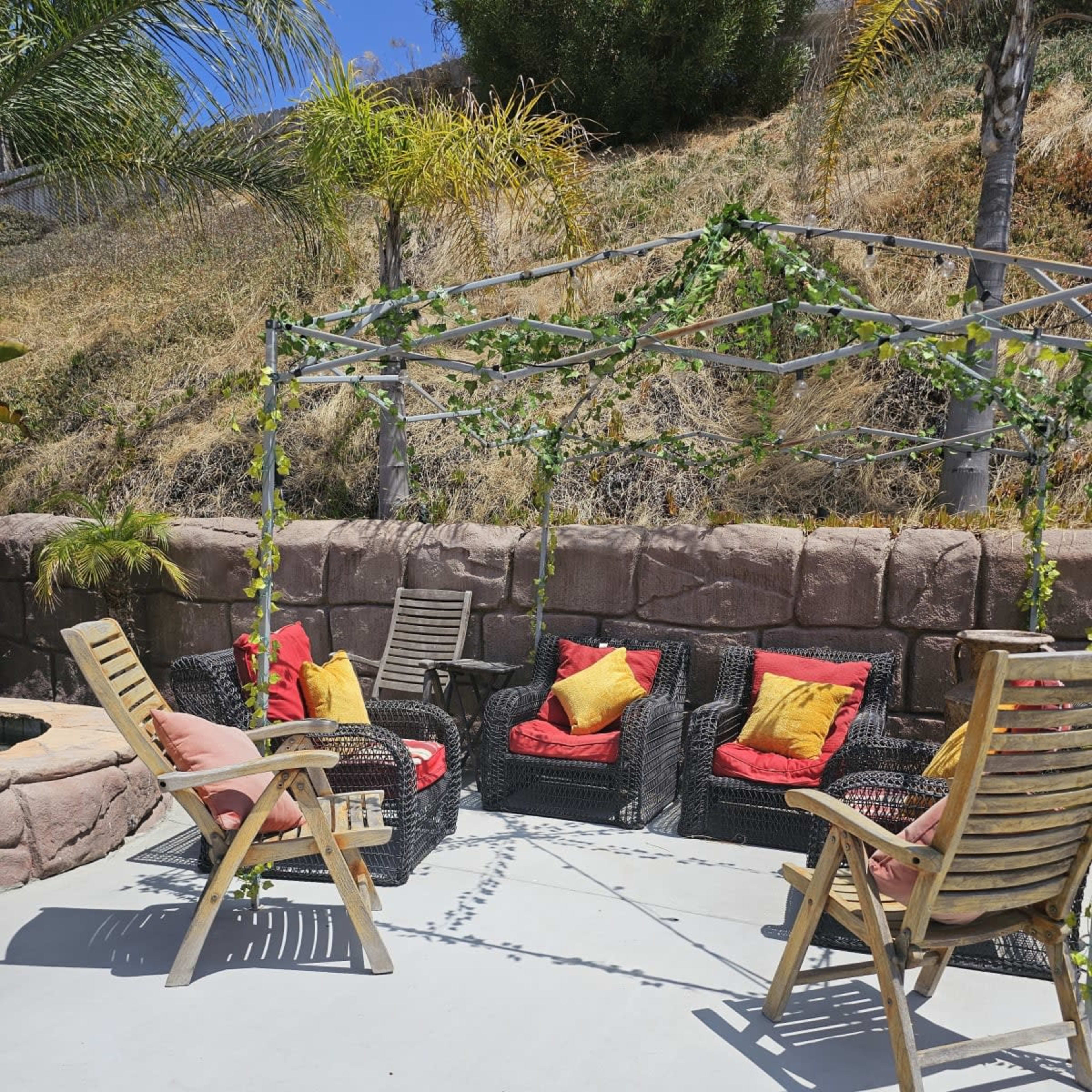 The scene features a modern outdoor seating area with two wooden chairs and a set of four cushioned chairs arranged under a vine-covered pergola.