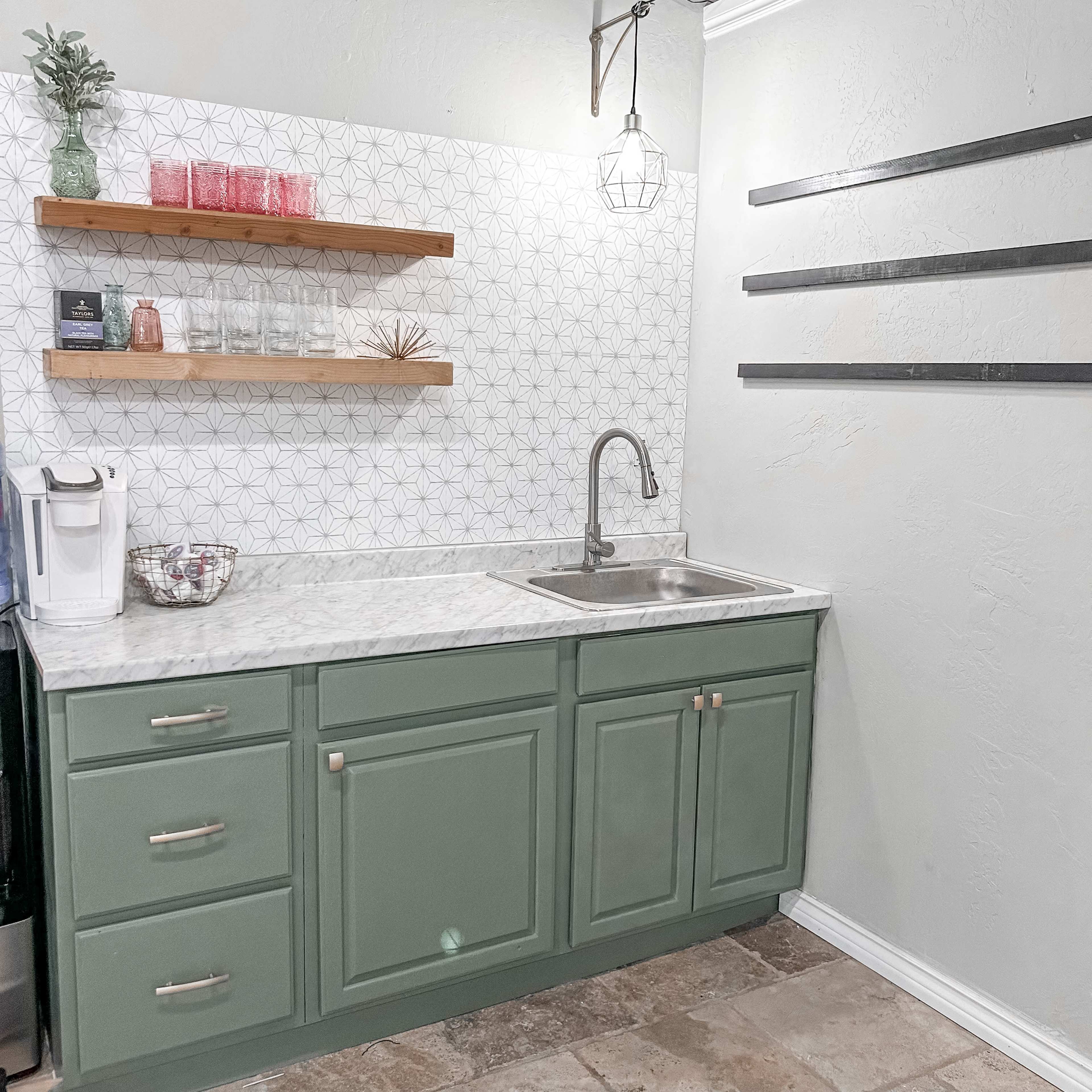 A modern kitchenette with green cabinetry, a stainless steel sink, a marble countertop, and wooden shelves displaying glassware and decorative items.