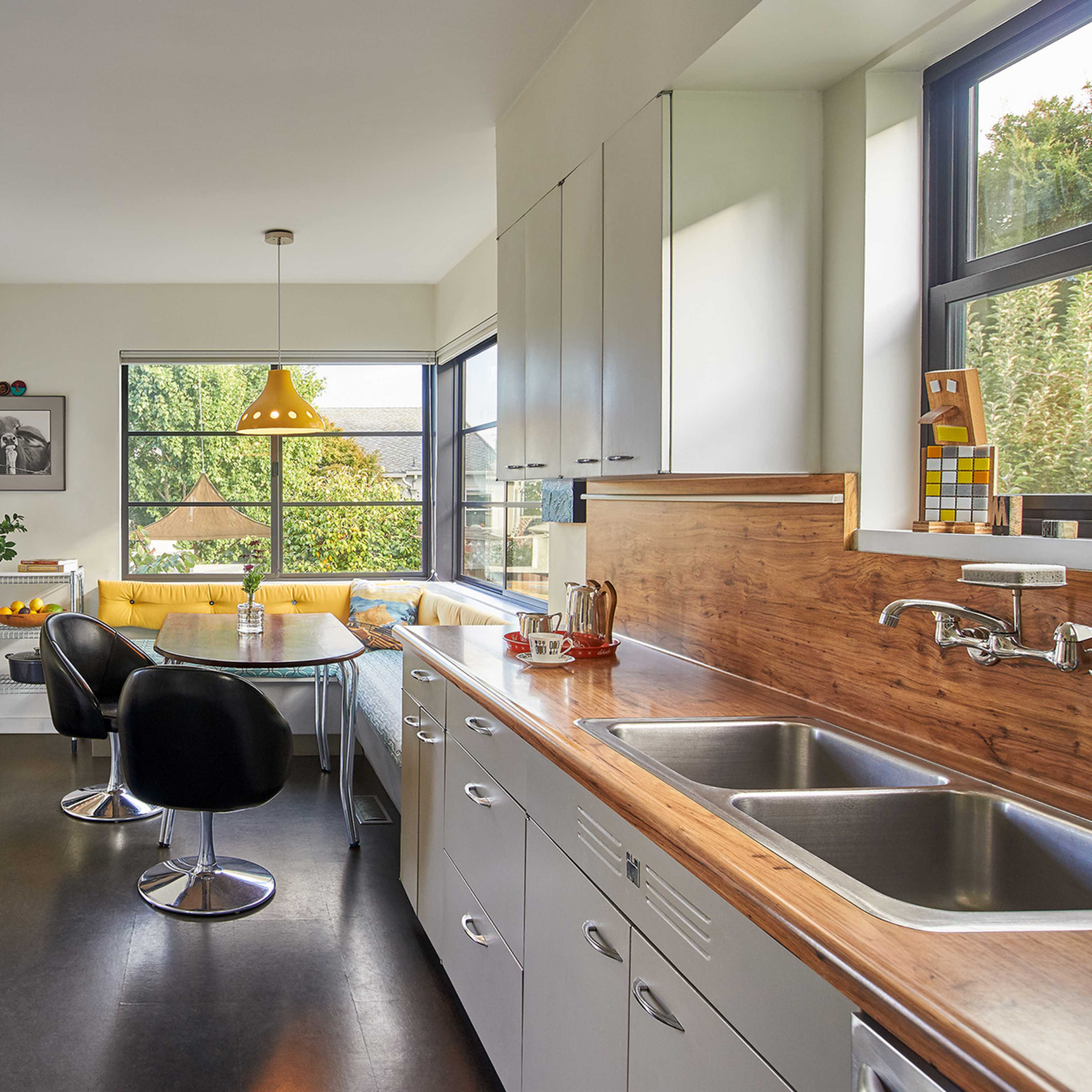A modern kitchen features a wooden countertop, stainless steel sinks, black chairs, and a dining area with a large window overlooking greenery.