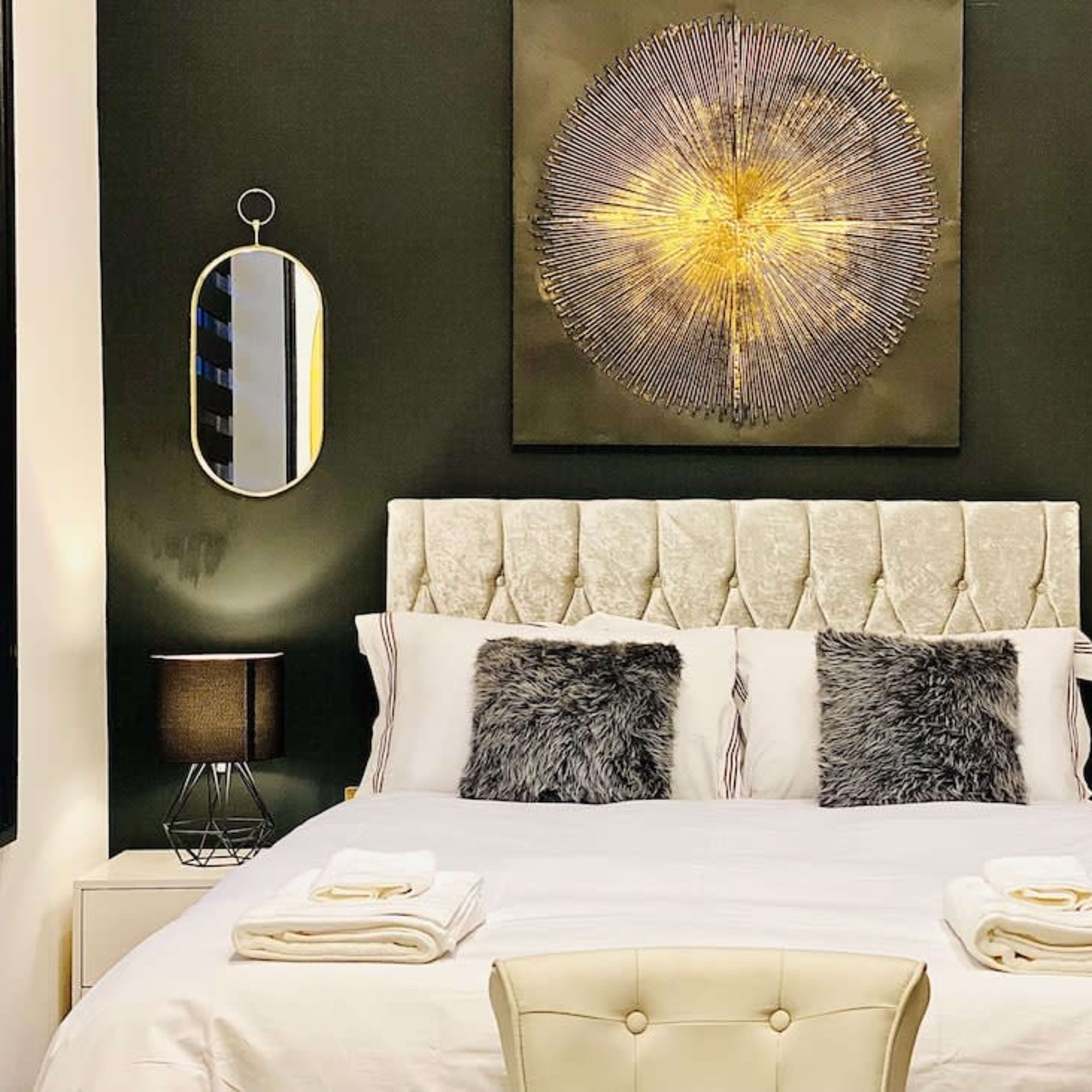 A neatly arranged bedroom with a tufted headboard, two bedside lamps, and decorative mirrors on dark walls.