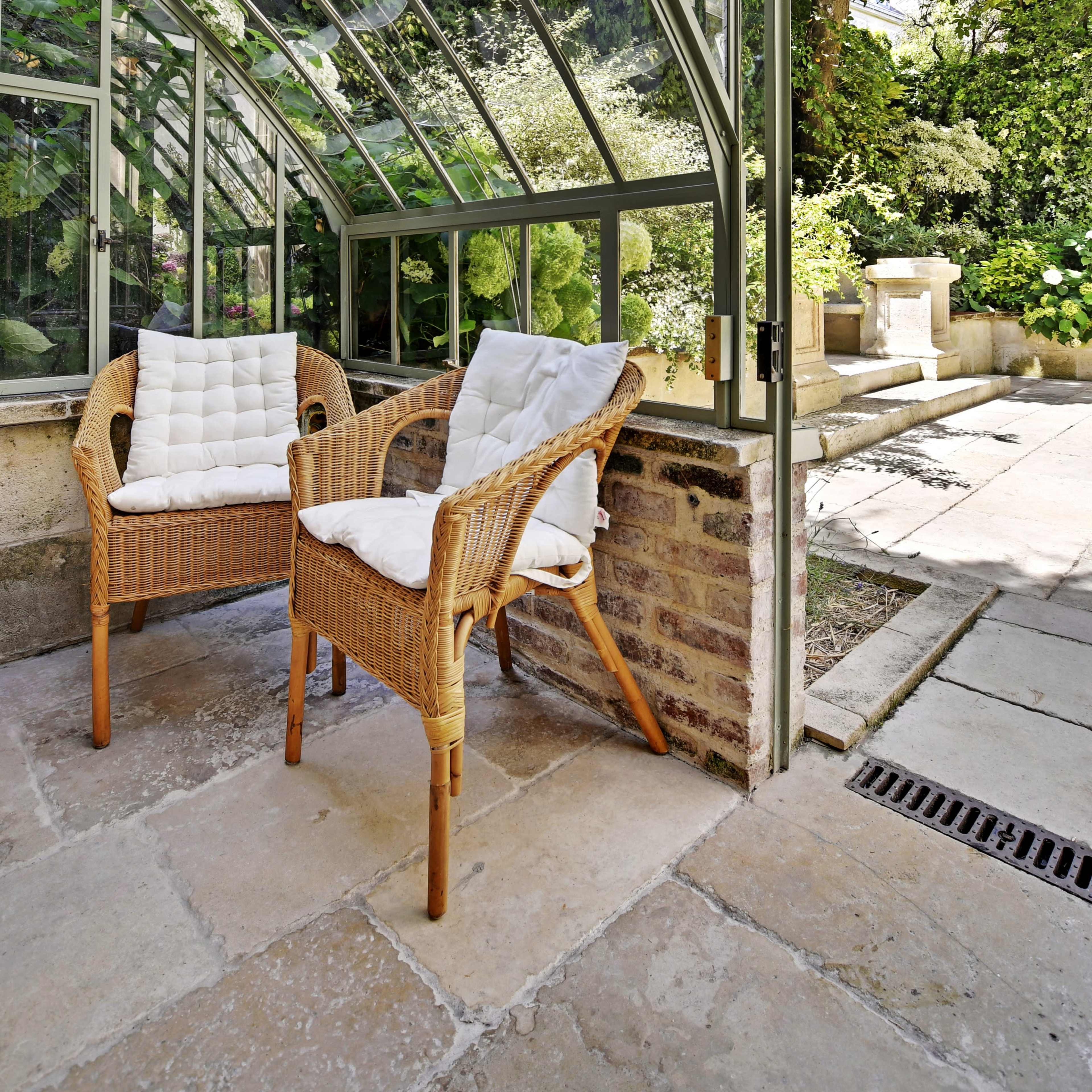 A conservatory features two wicker chairs with cushions, surrounded by lush greenery and stone flooring.