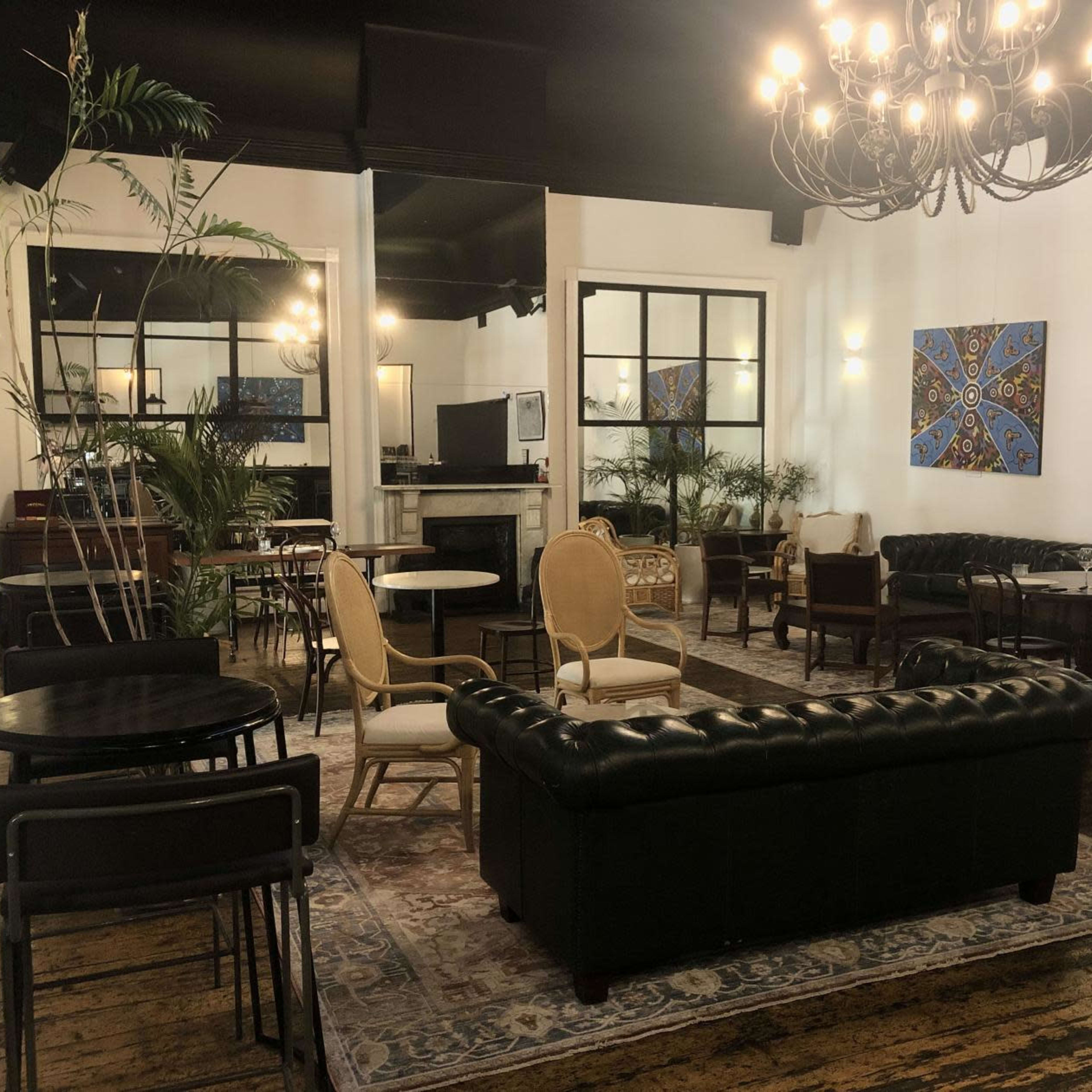 A stylish lounge area with various seating arrangements, including a leather sofa, wooden tables, and potted plants, set against a backdrop of white walls and artwork.