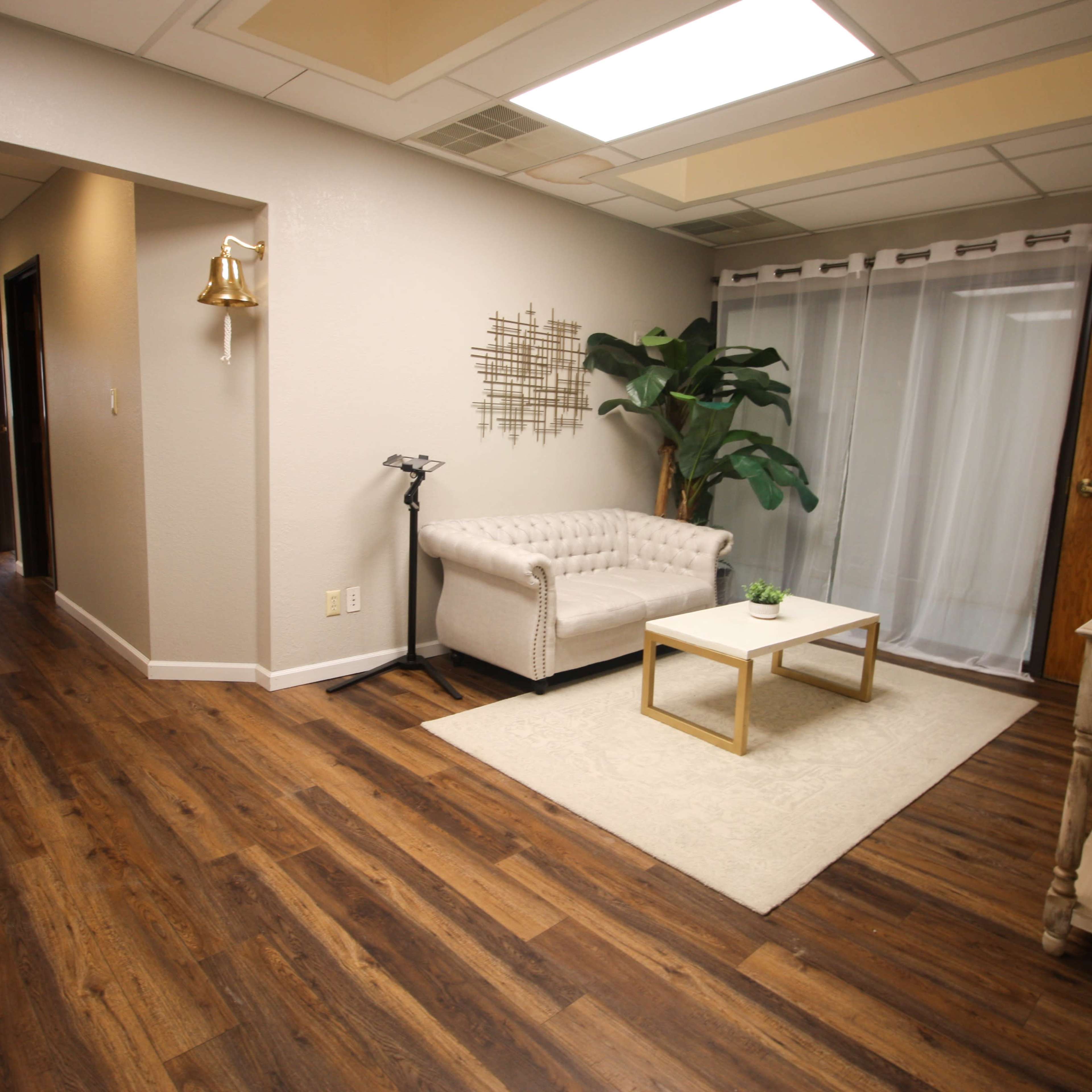 A well-lit waiting area features a white tufted sofa, a small coffee table, a potted plant, and wooden flooring.