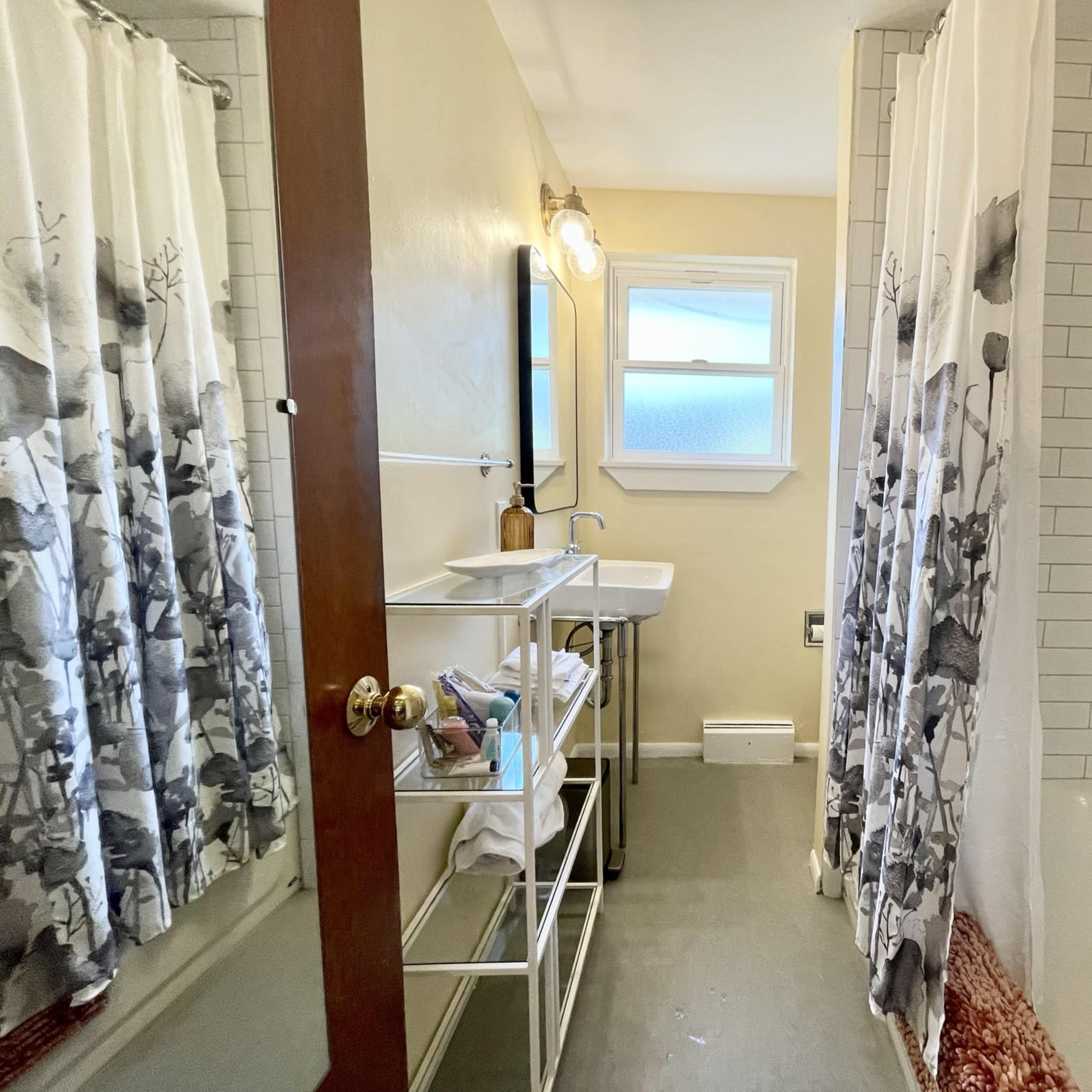 A bathroom with a shower area, a bathtub, a sink on a metal stand, and light-colored walls.