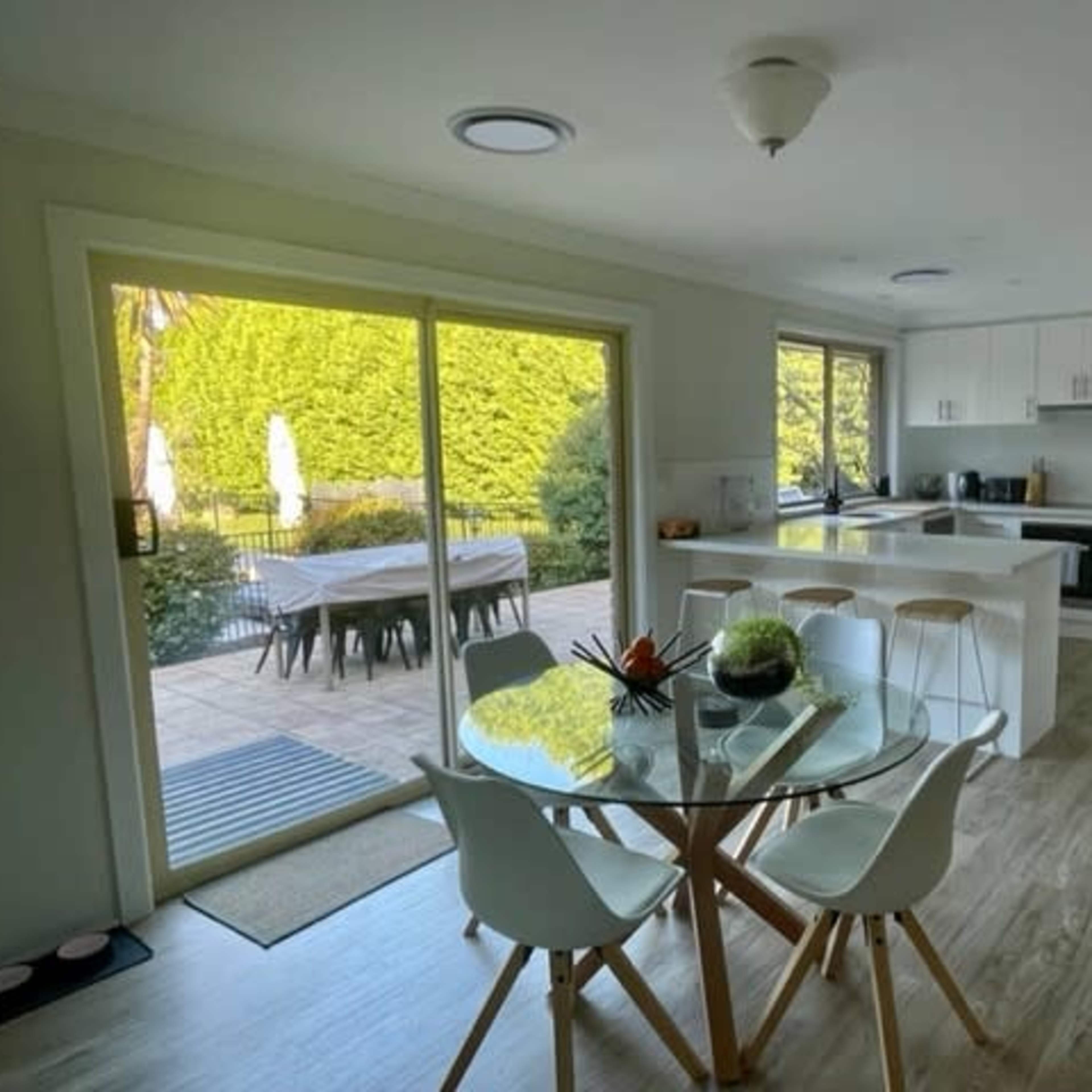A modern kitchen and dining area features a round glass table with white chairs, overlooking a patio and garden through large sliding doors.