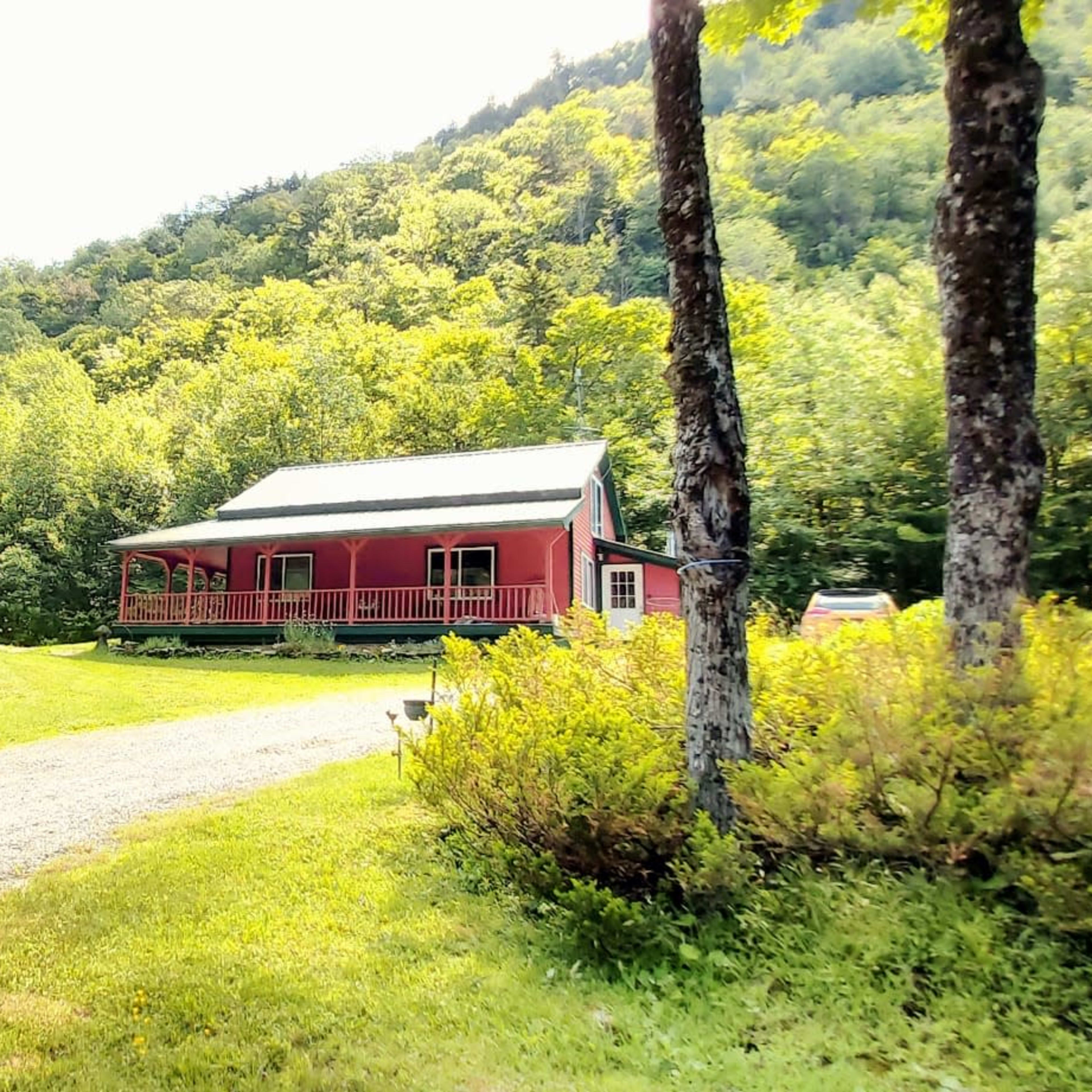 A red house with a porch is located beside a gravel driveway amidst green trees and a hillside.