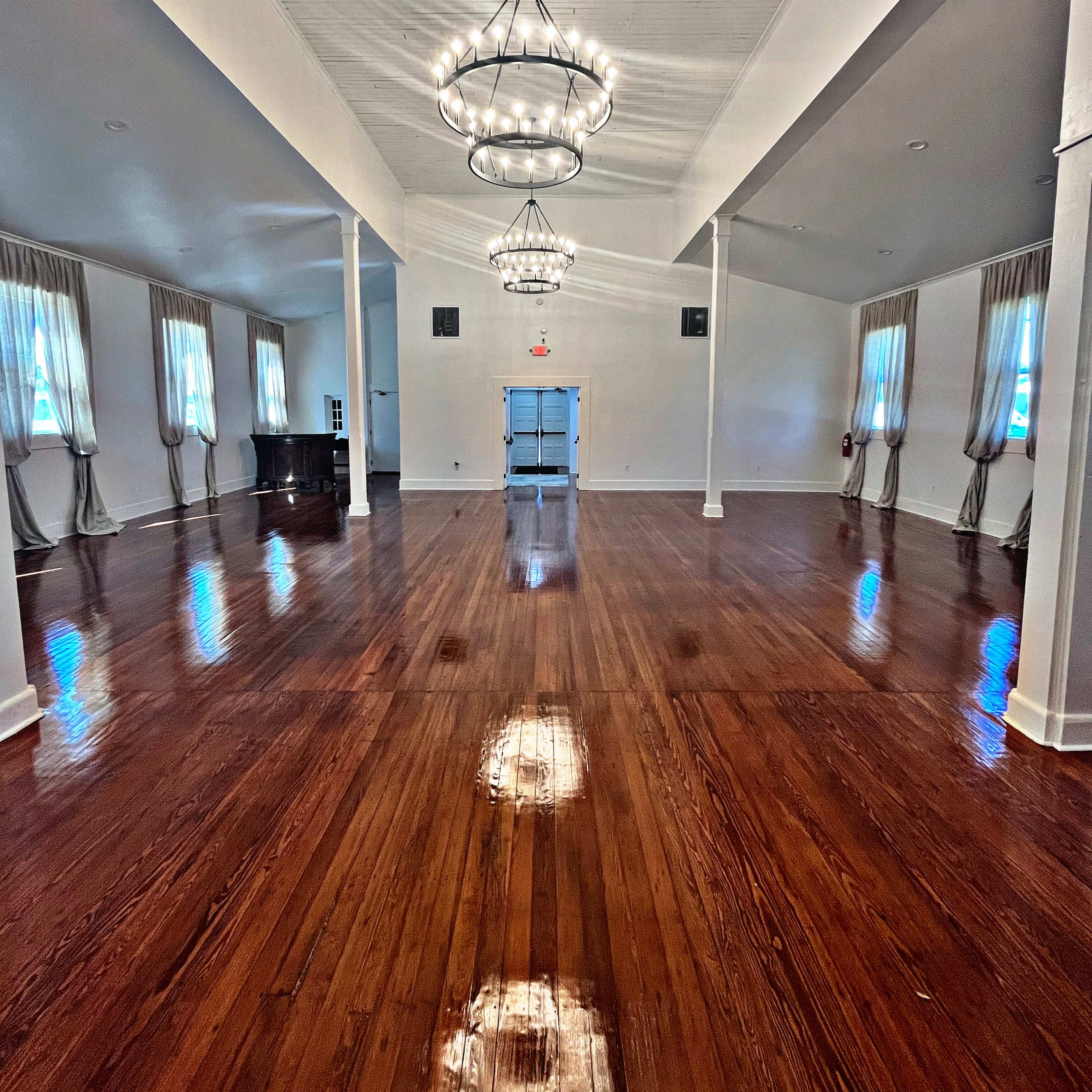A spacious, empty hall with polished wooden floors, large windows with drapes, and modern chandeliers hanging from the ceiling.