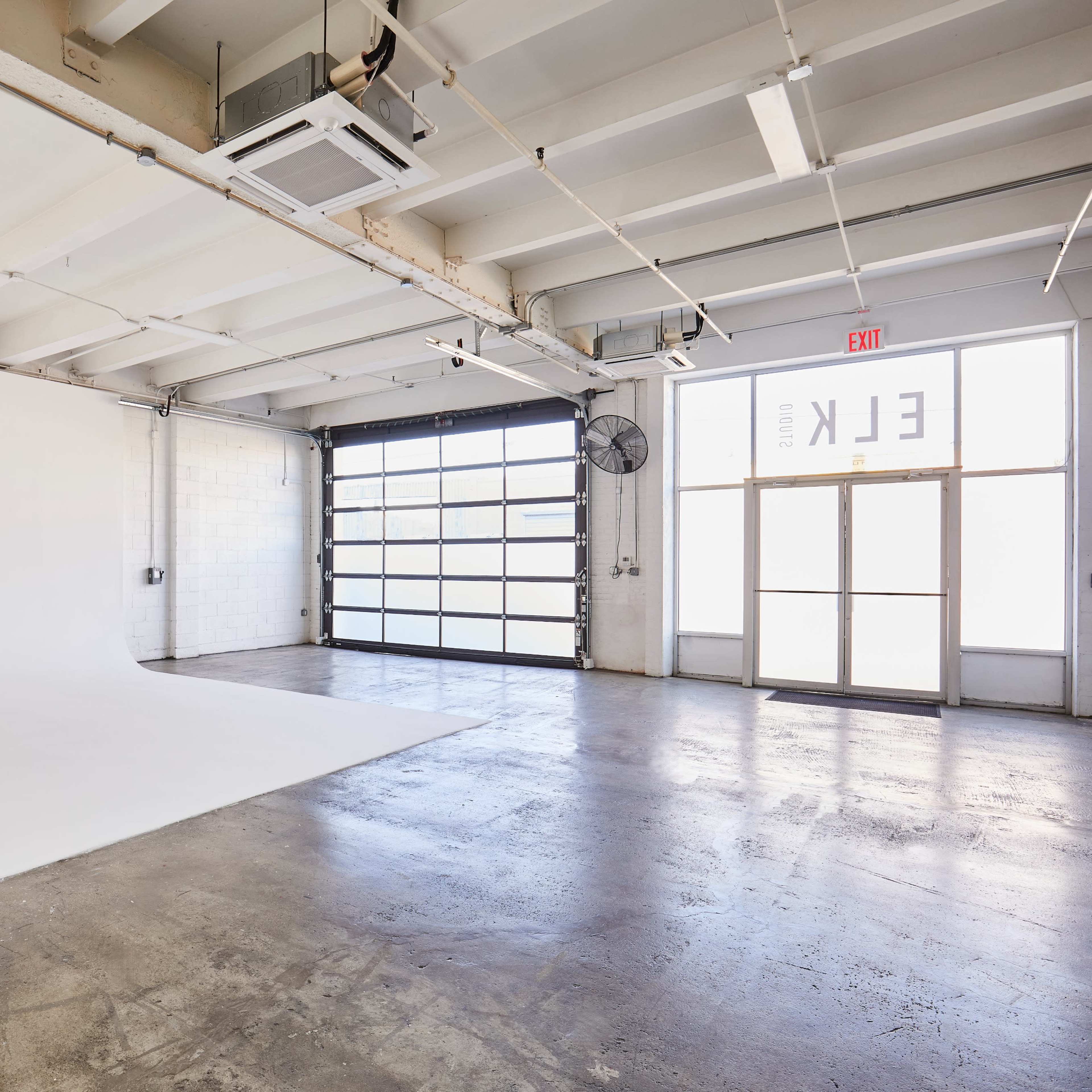 A spacious studio with a white backdrop and large windows, featuring a concrete floor and exposed beams.