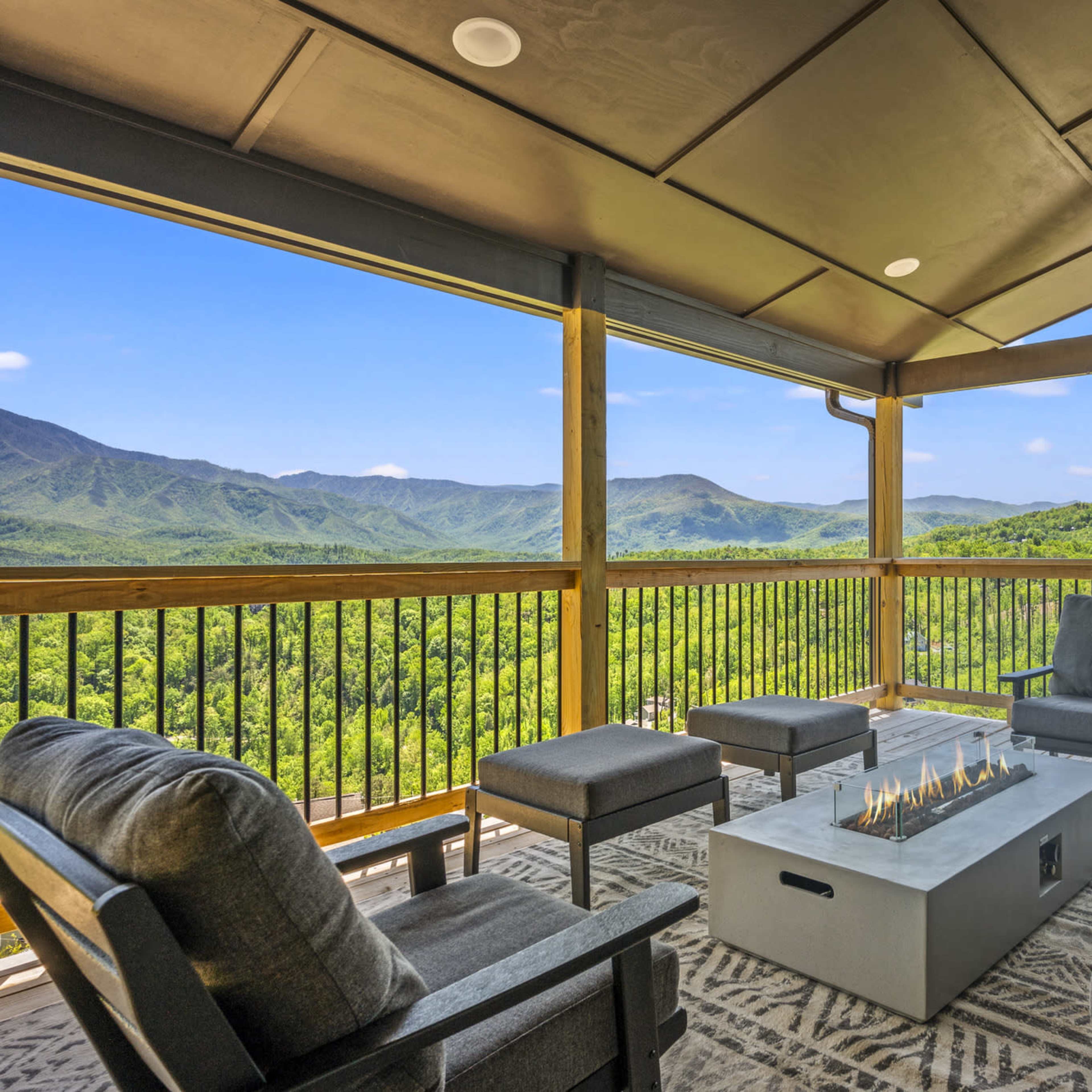 A covered balcony features comfortable seating and a modern fire pit, overlooking a lush mountain landscape.