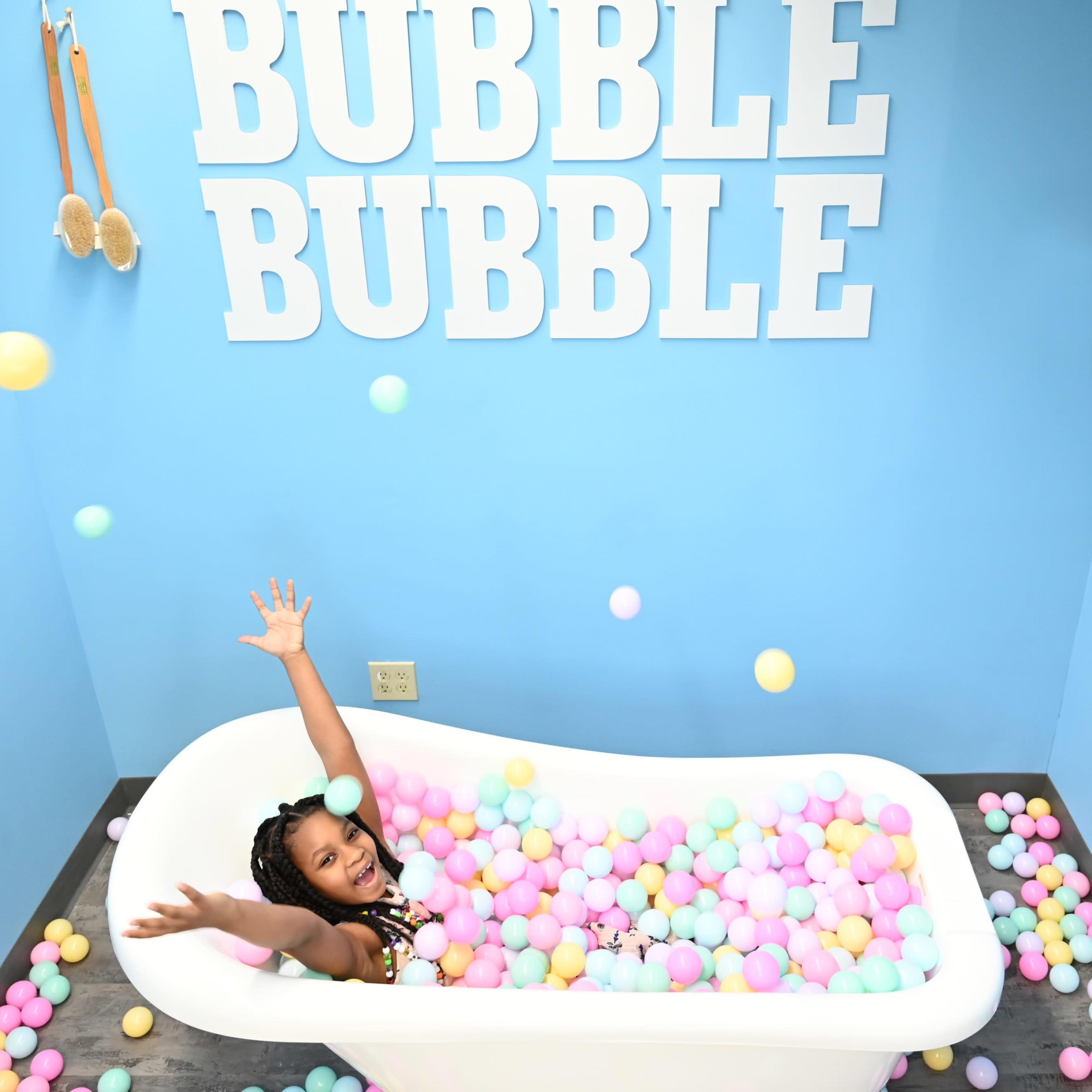 A girl sits in a white bathtub filled with colorful balls, surrounded by blue walls with the words "BUBBLE BUBBLE" displayed above her.