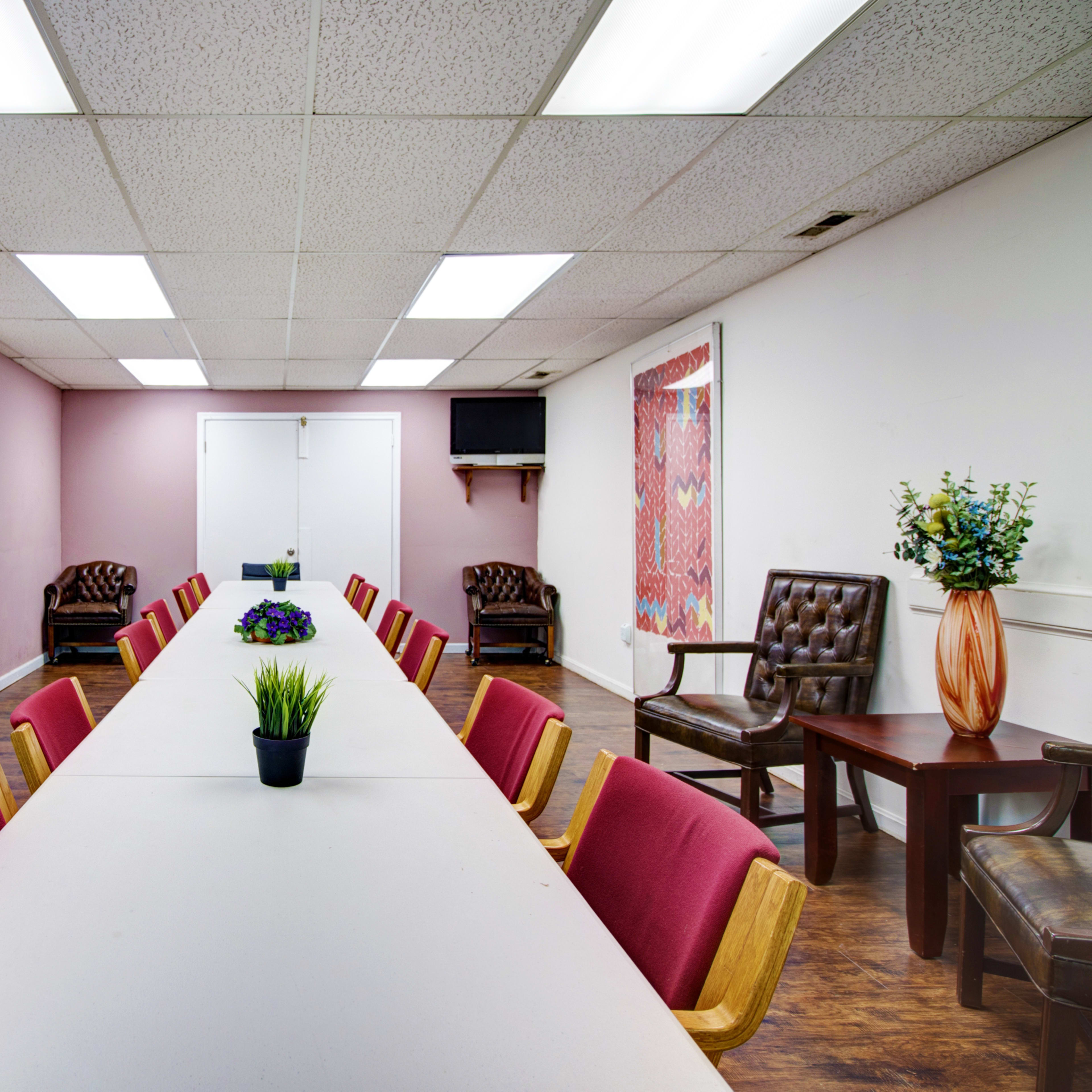 A meeting room with a long table surrounded by chairs, flanked by two doors and a wall-mounted TV.