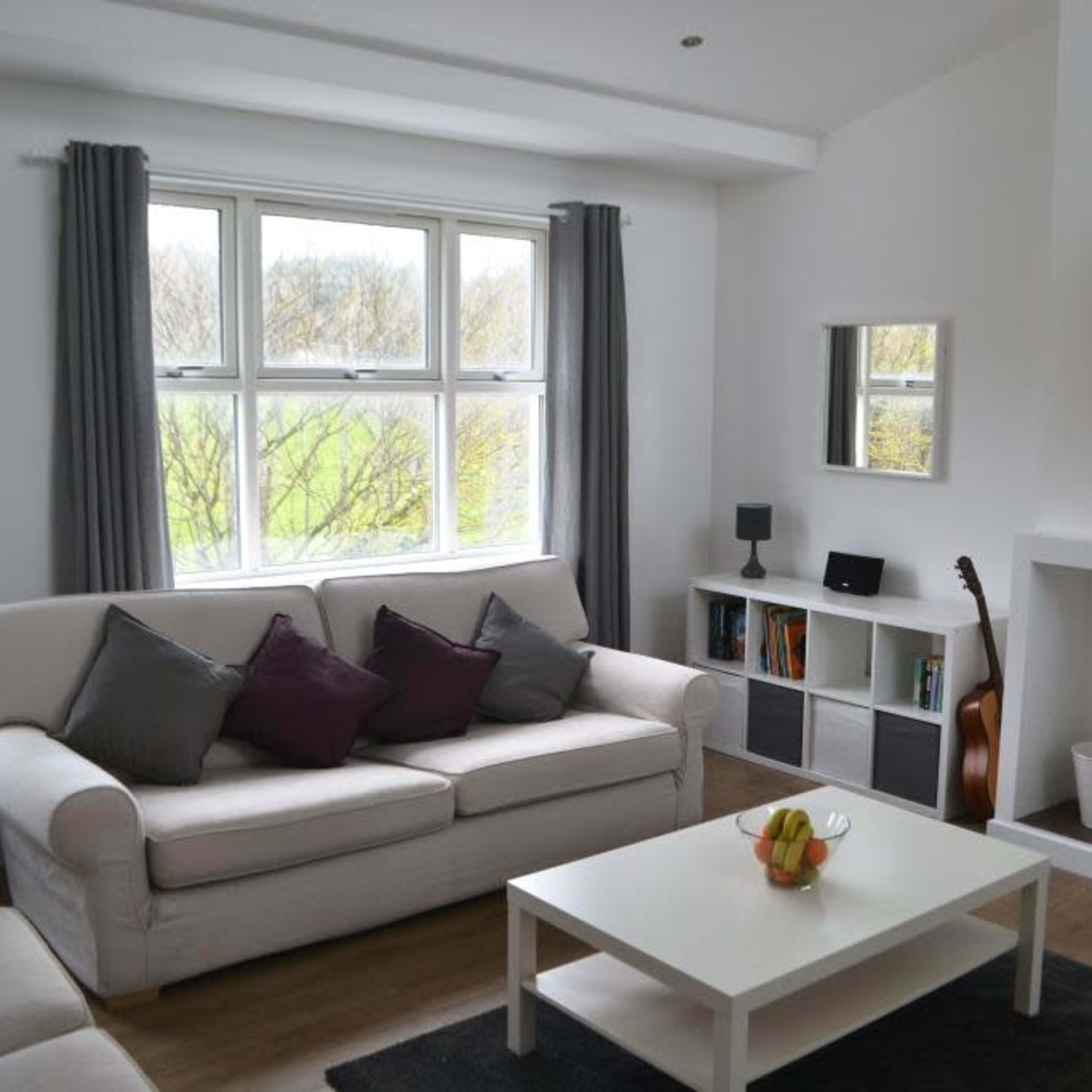 A modern living room features a beige sofa, a white coffee table with a fruit bowl, a wall-mounted TV, and a window with grey curtains overlooking greenery.
