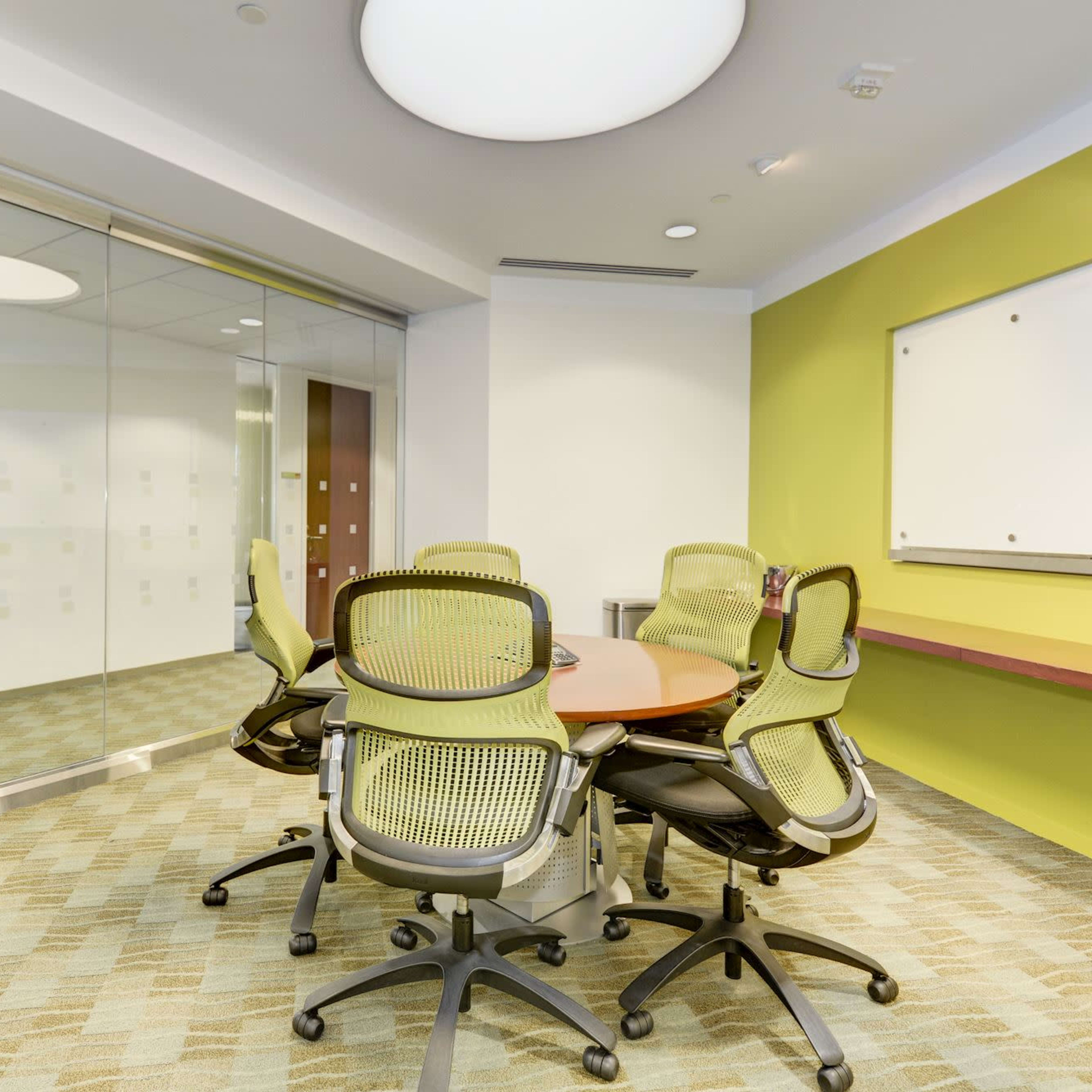 A conference room features a round table with six green office chairs, surrounded by a whiteboard and glass walls.