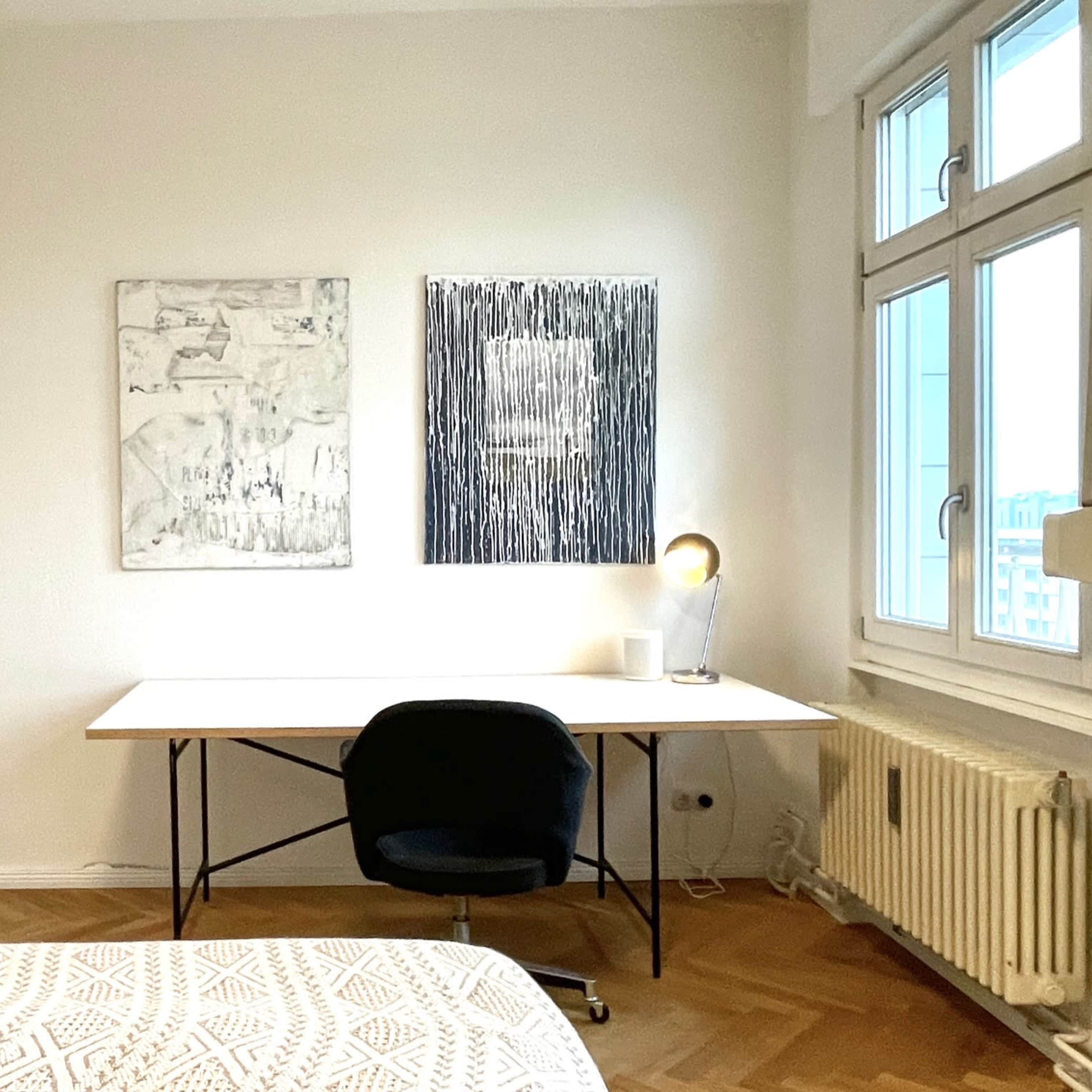 A minimalistic bedroom featuring a bed, a desk with a lamp, and two framed artworks on the wall.