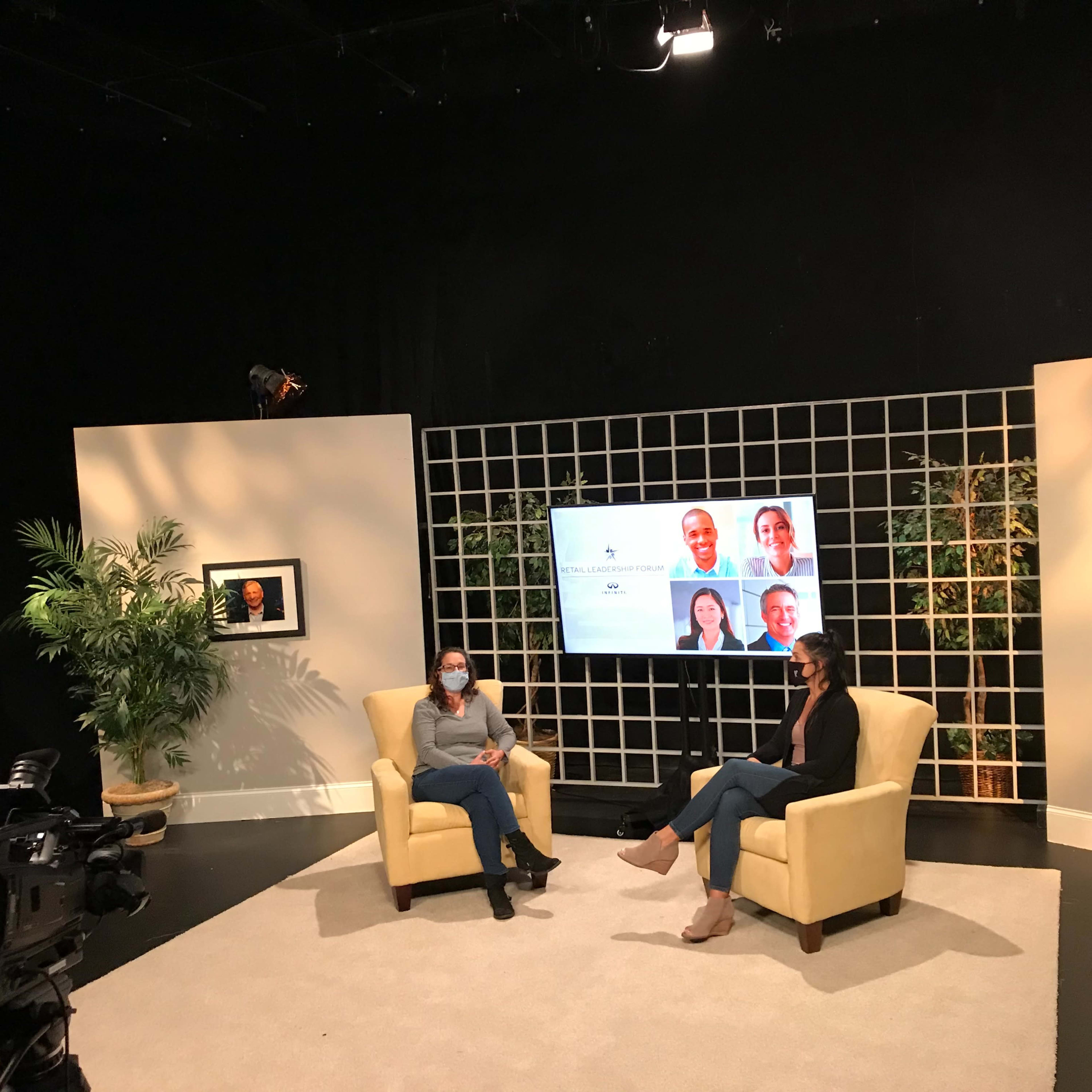 Two women are seated in a television studio set, facing each other, with a large screen displaying headshots in the background and cameras positioned to capture the scene.