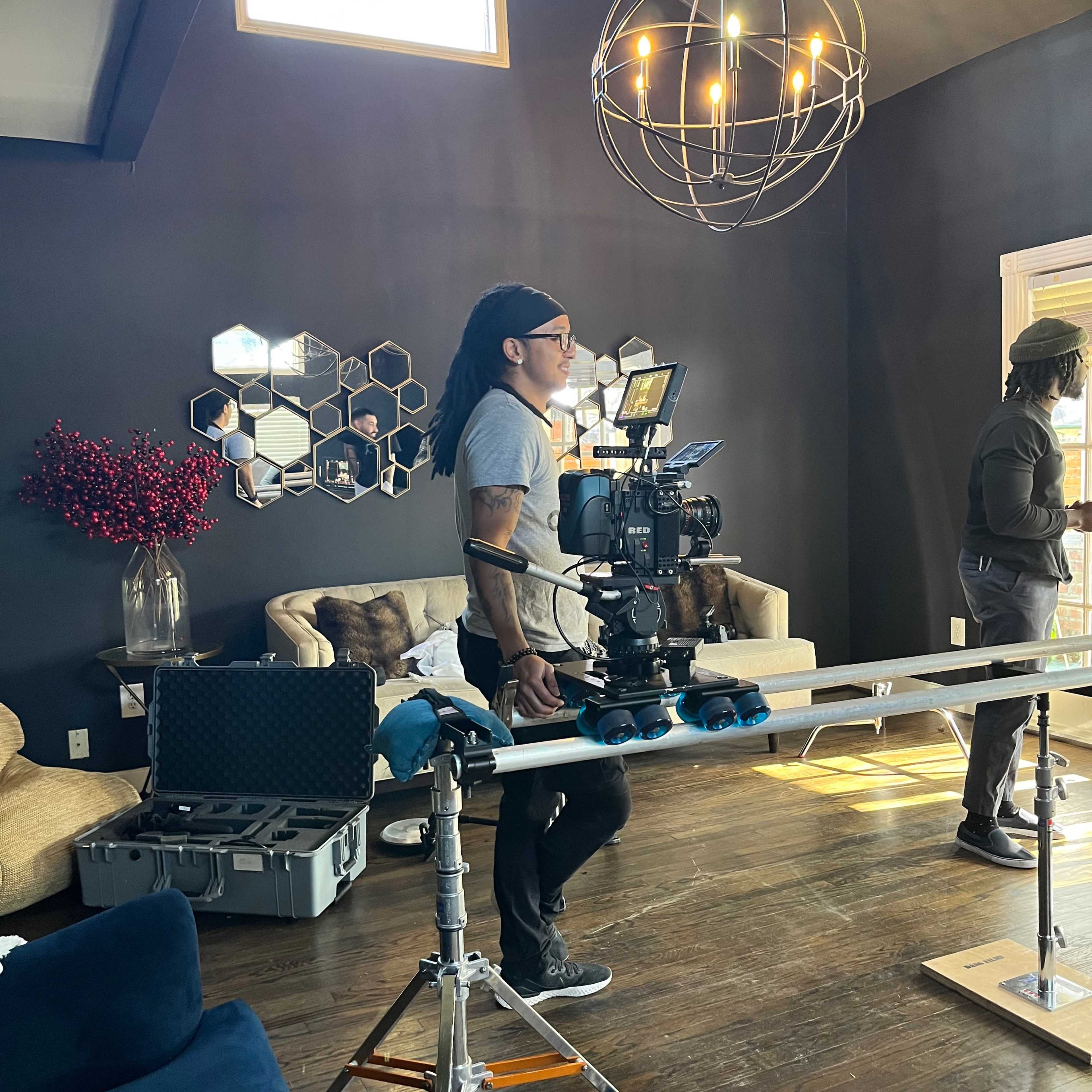 A man operates a camera on a track system in a well-furnished living room with large windows and decorative mirrors.