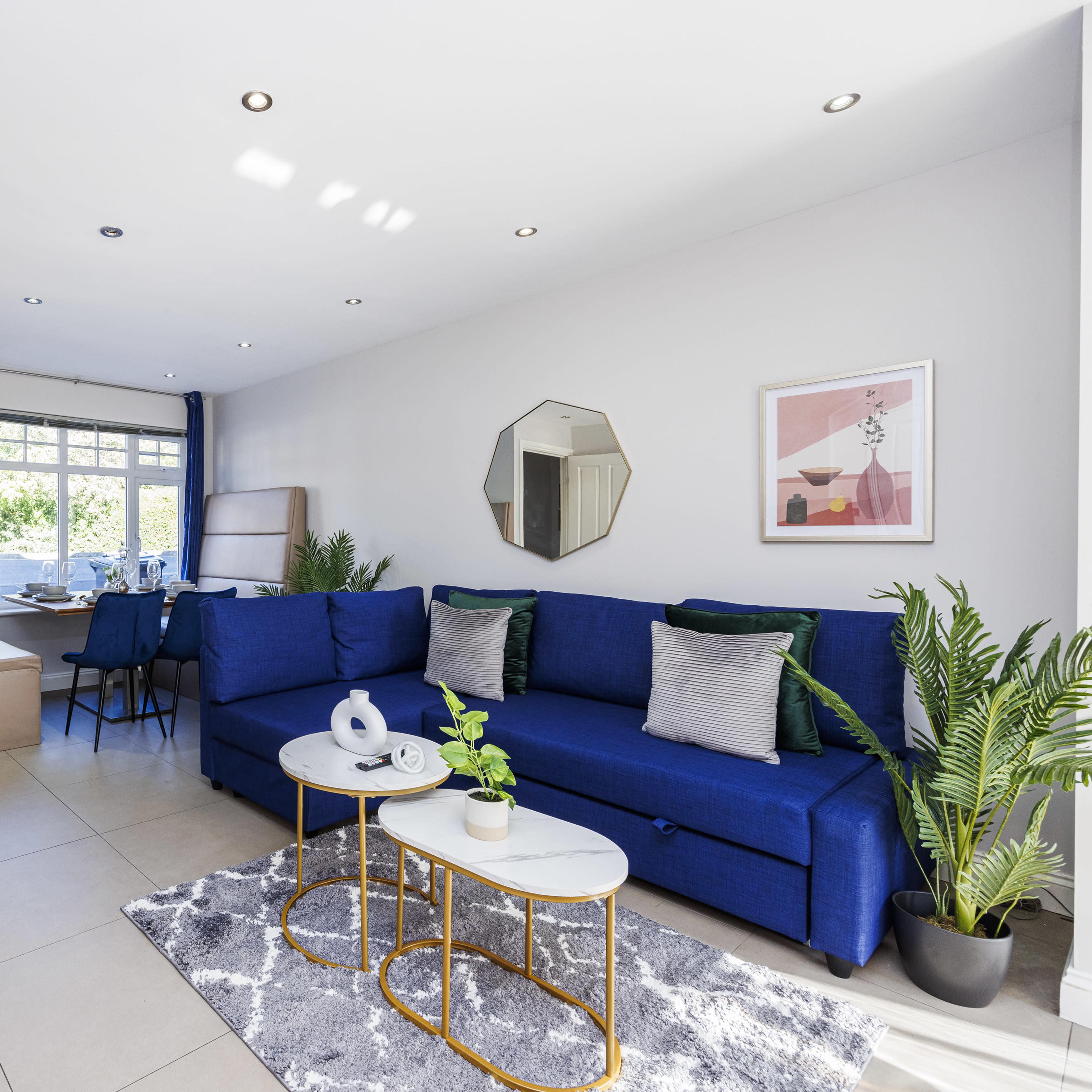 A modern living room with a blue sectional sofa, a coffee table, green plants, a television, and a dining area visible in the background.