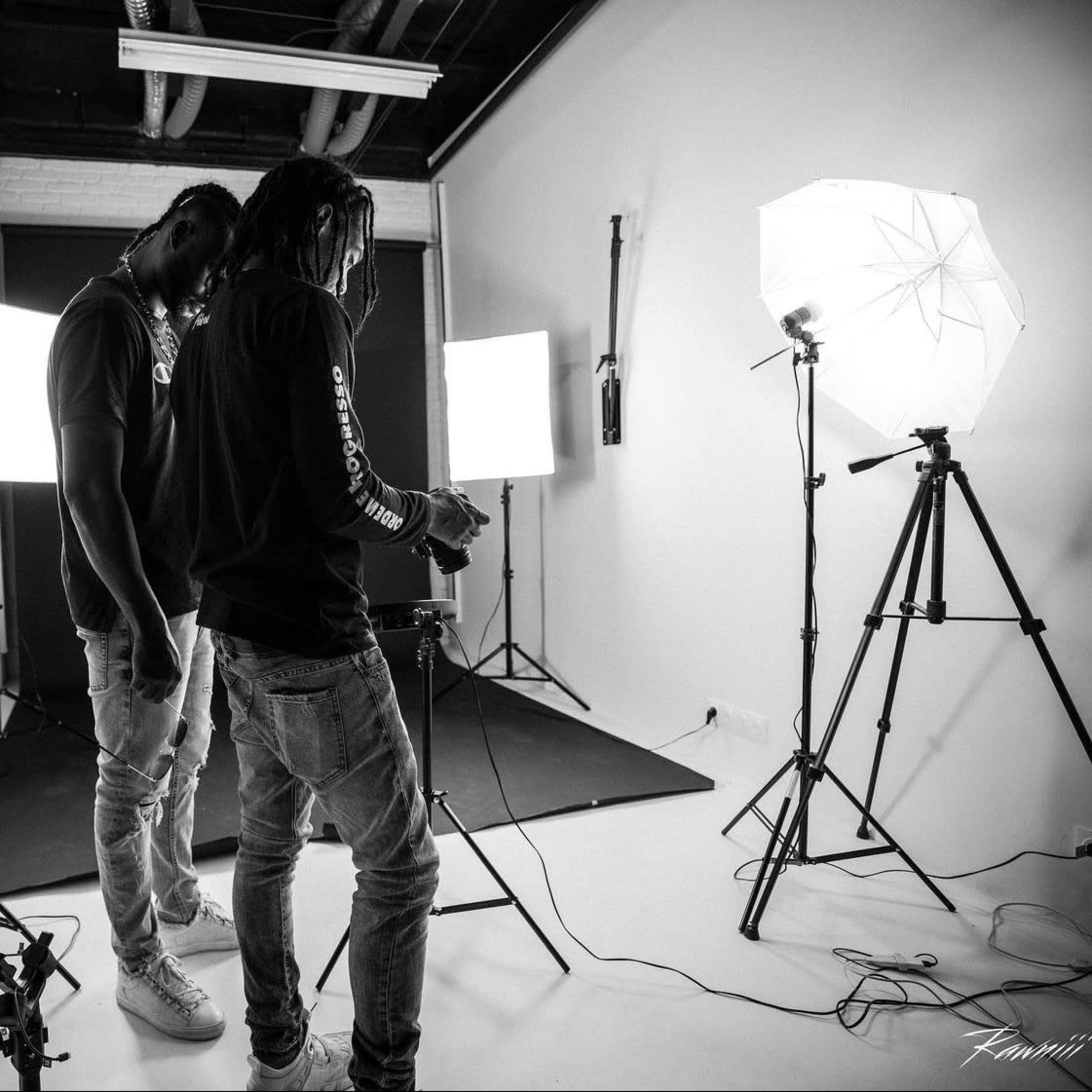 Two individuals set up photography equipment in a studio with softbox lights and a camera on a tripod.