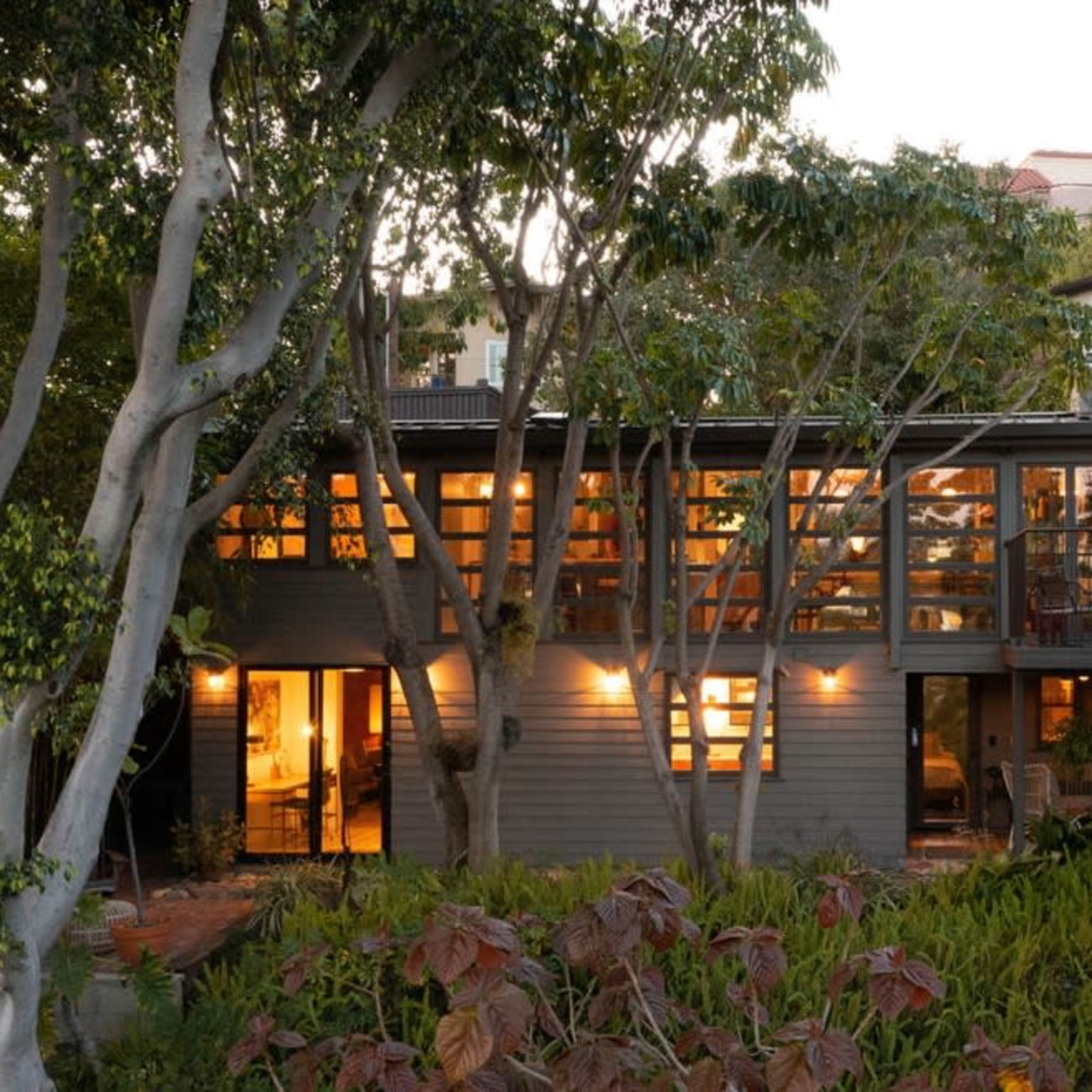 A modern two-story house is surrounded by trees and gardens, with warm lights glowing from its large windows at dusk.