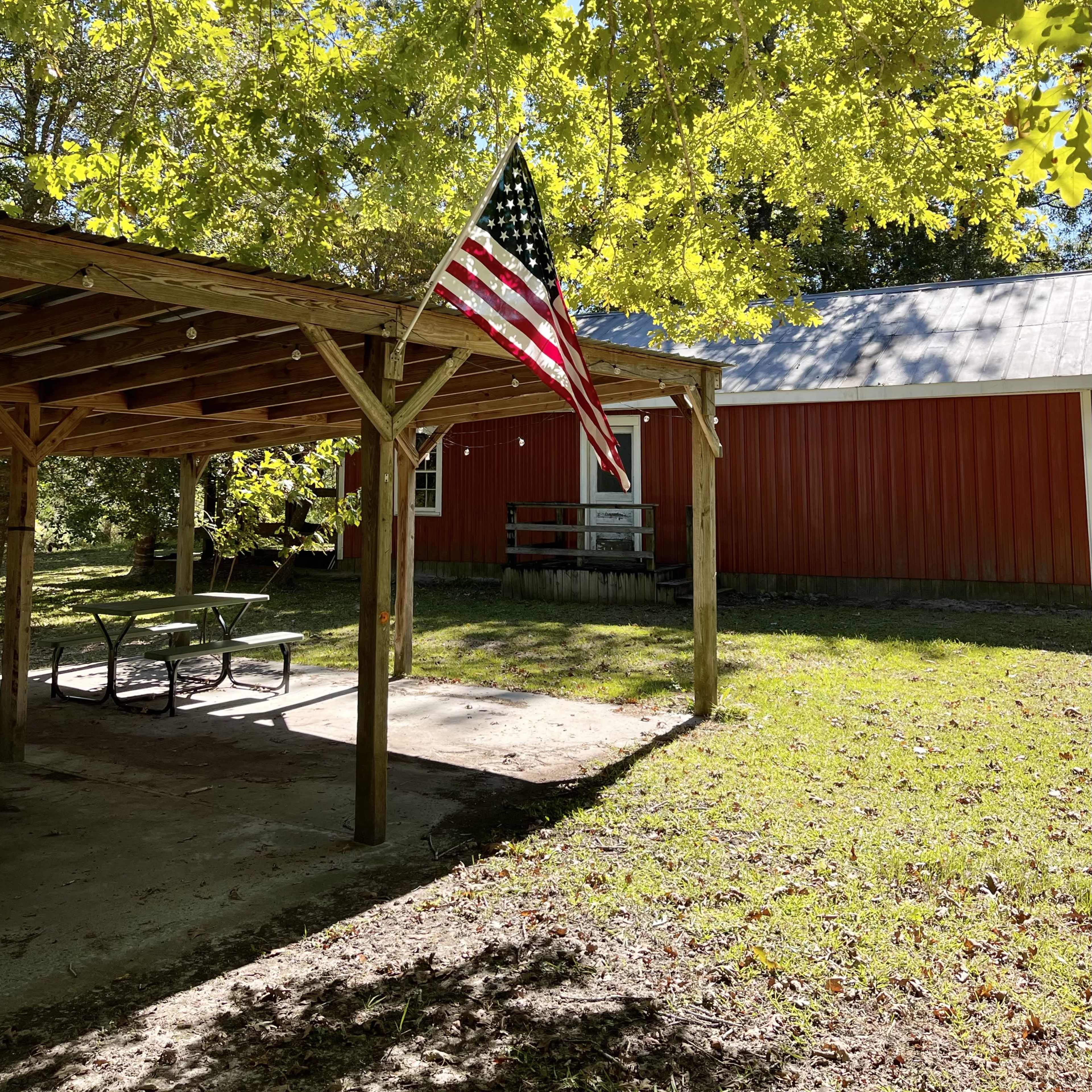 A red building with a white roof is partially visible behind a covered structure, under which an American flag hangs amid green trees.