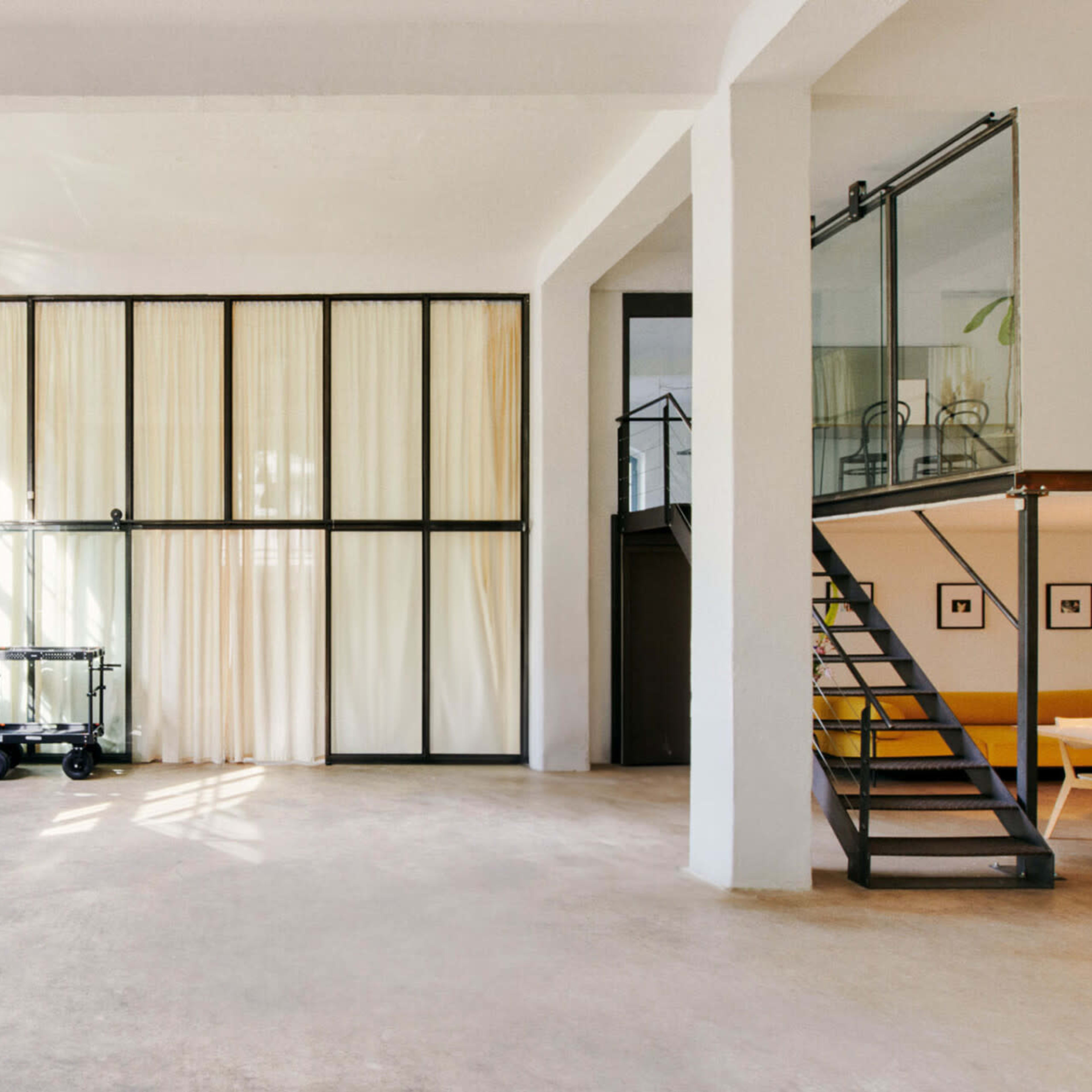 A spacious, well-lit room features large windows, a curtain wall, and a staircase leading to a loft area.