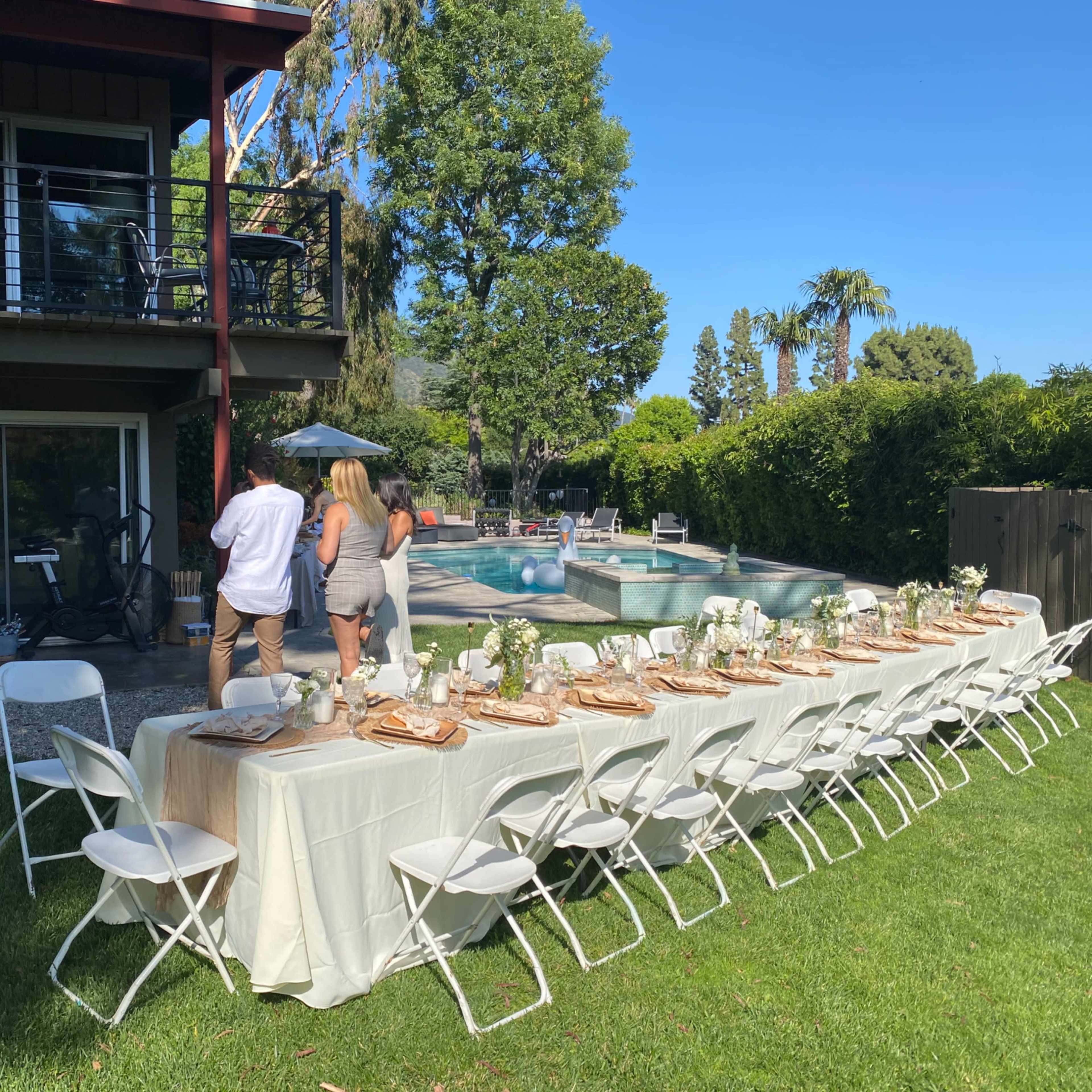 A long dining table is set up outdoors near a pool, with guests walking towards it while surrounded by greenery and a two-story house in the background.