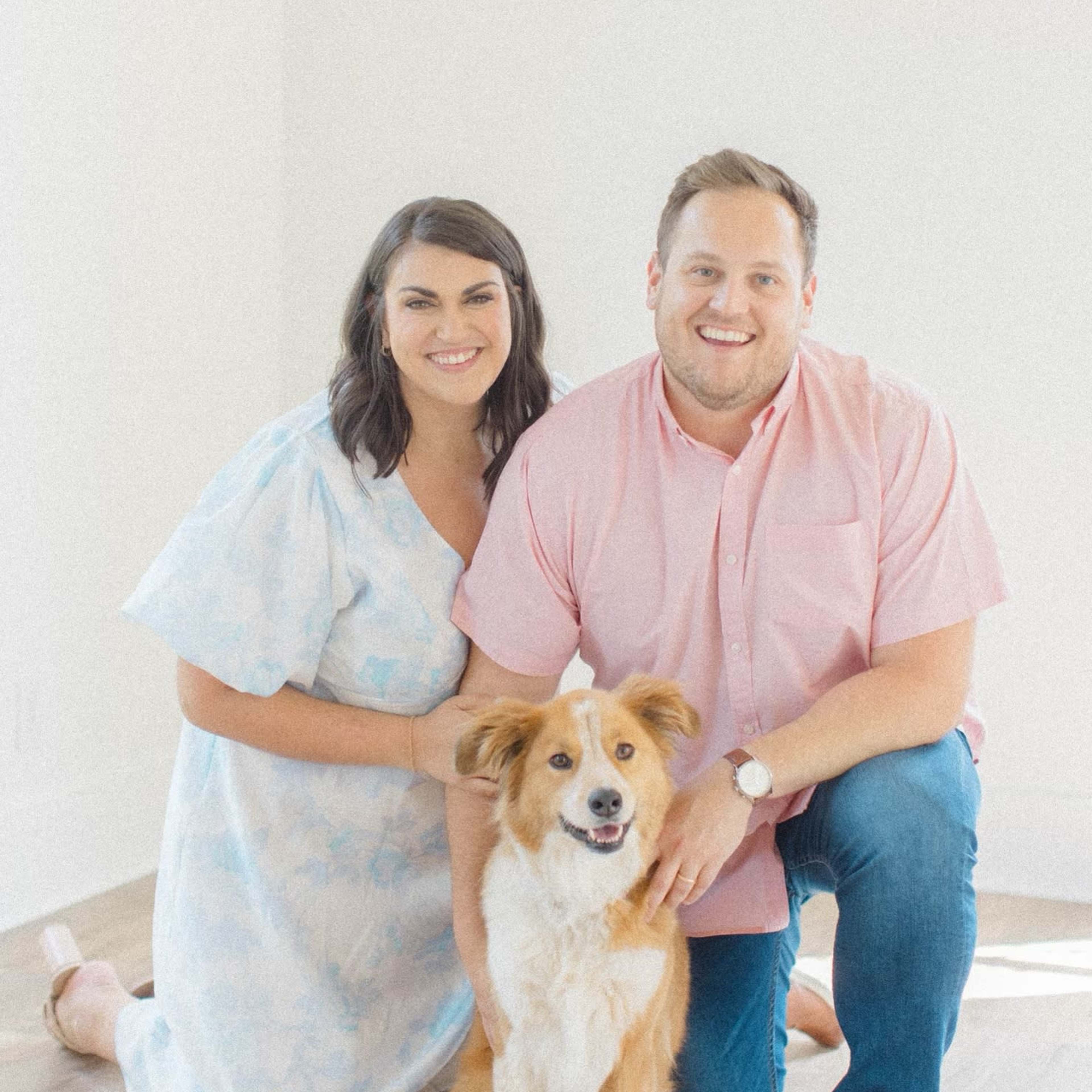 A couple is kneeling on a light-colored floor with a dog in front of them, smiling in a well-lit room.