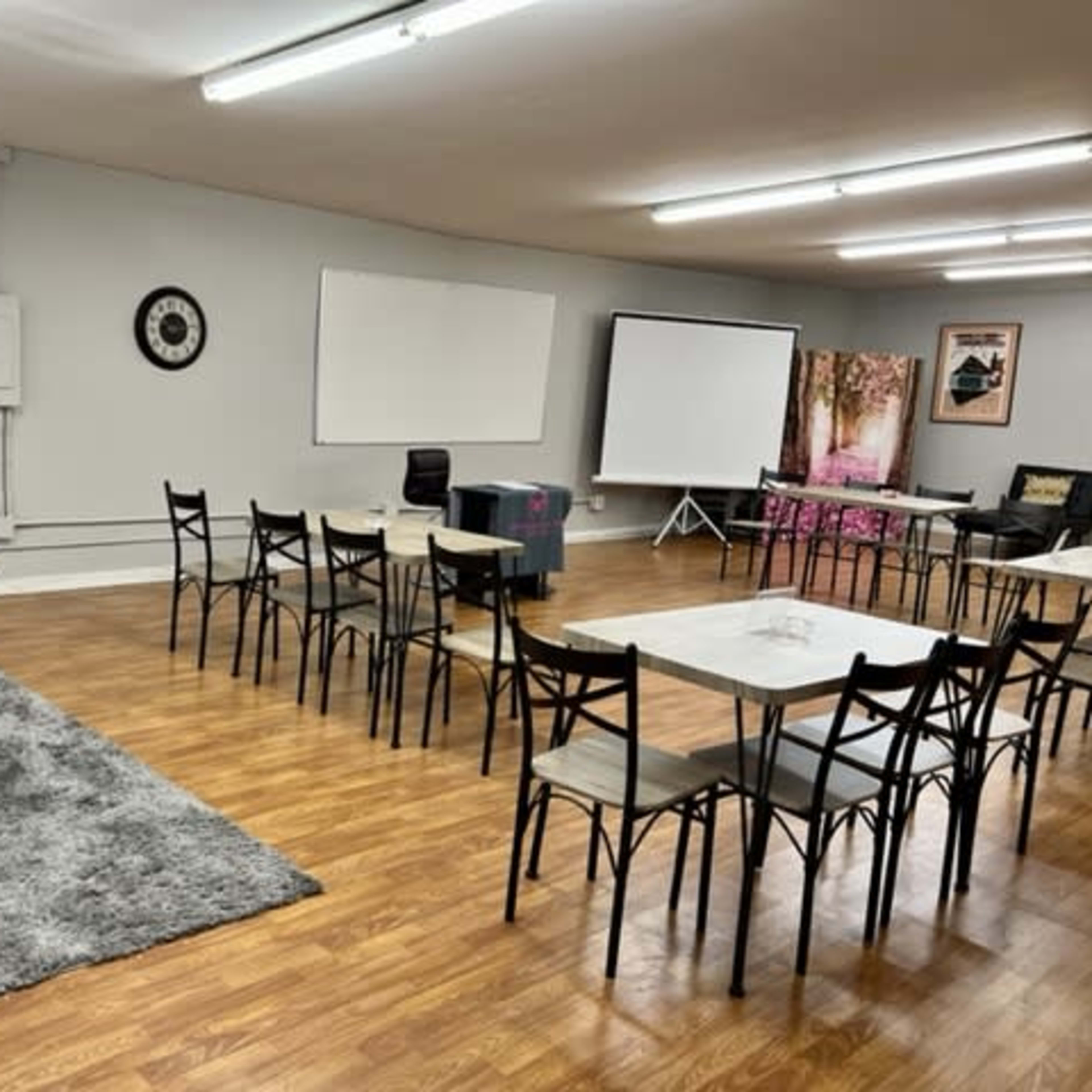 A spacious meeting room features a series of tables and chairs arranged in a circle, with a projector screen and whiteboards on opposite walls.
