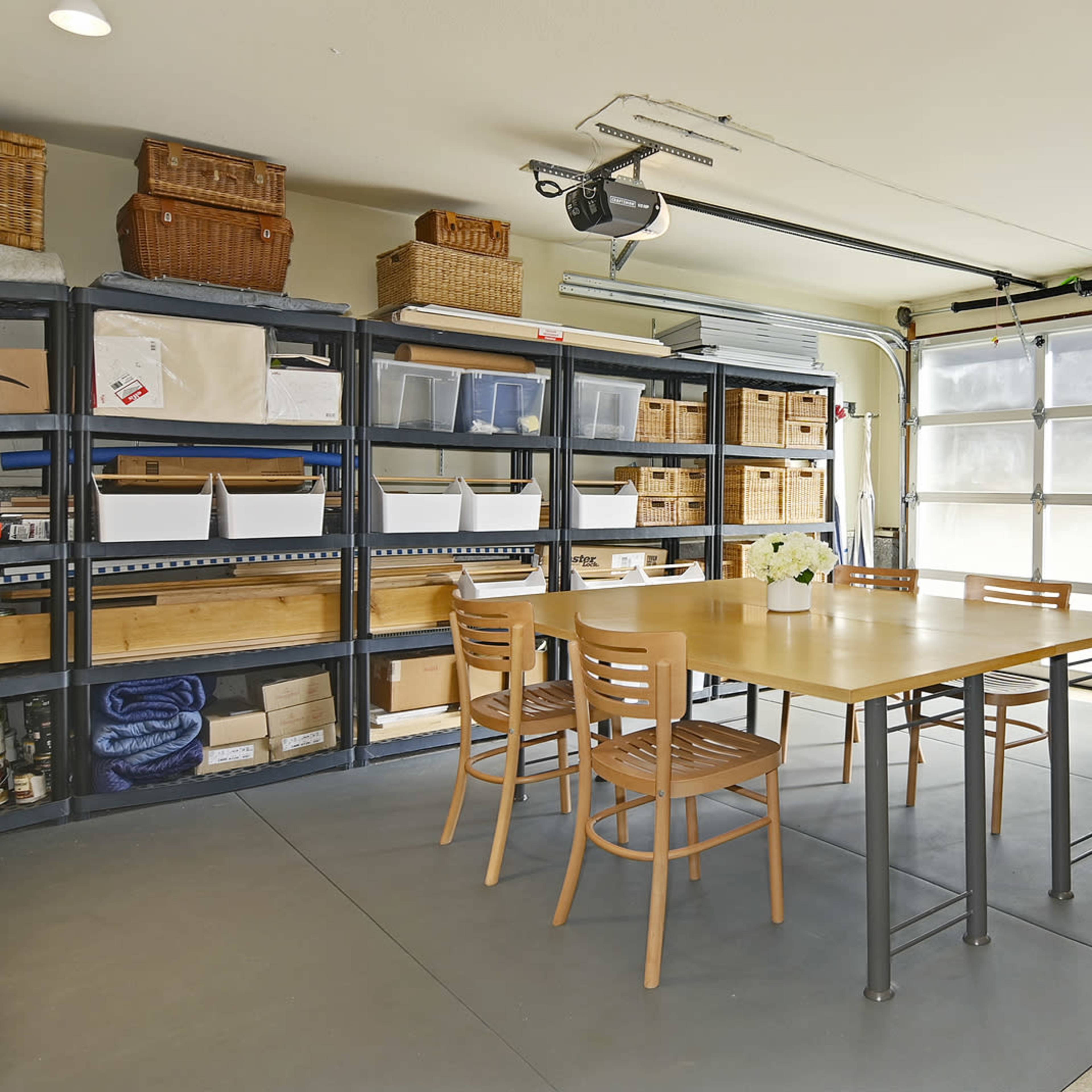 A well-organized garage features a central table with four chairs, surrounded by shelves filled with baskets and storage boxes, and a large garage door.