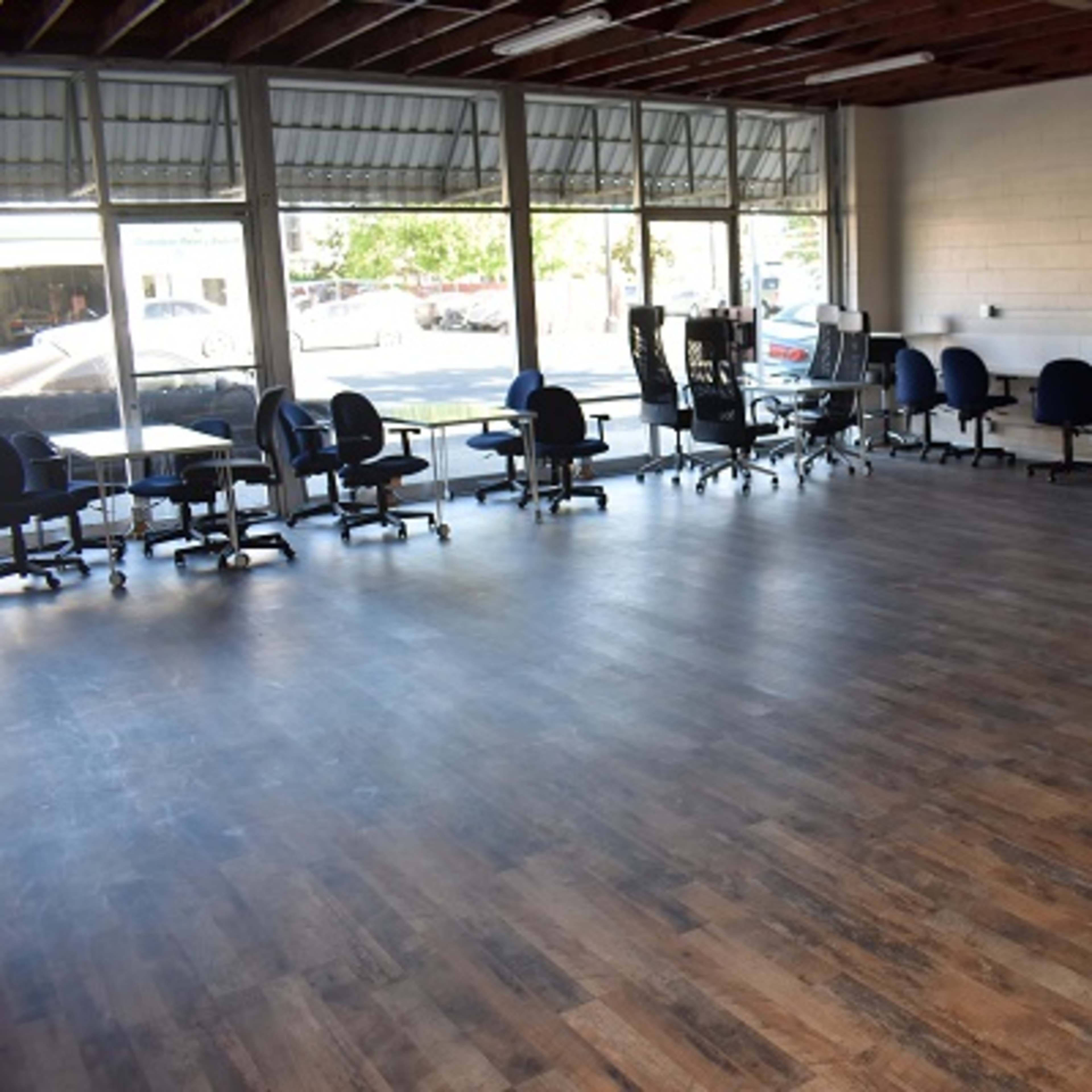 An empty room with wooden flooring, lined with office chairs and tables, and large windows letting in natural light.