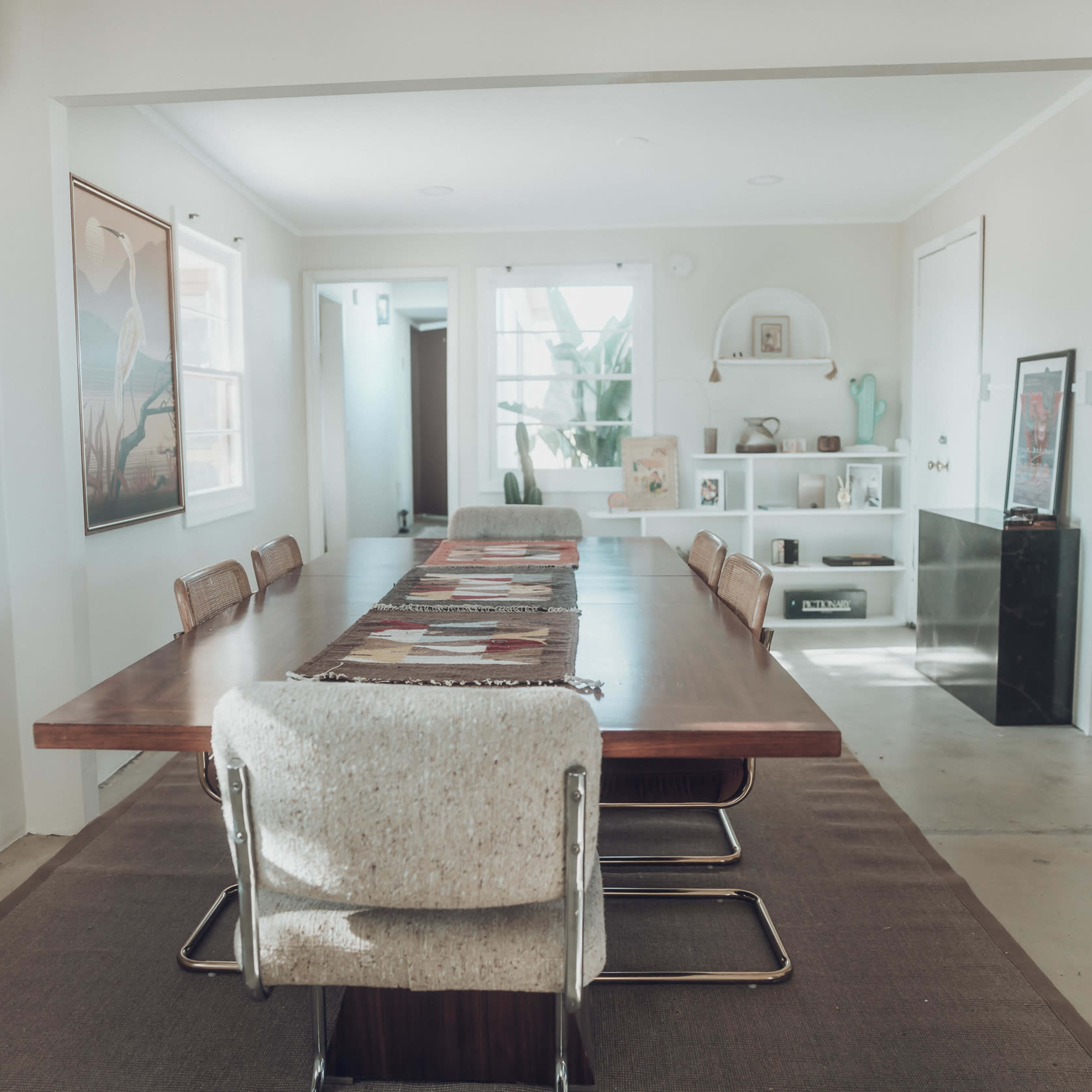 A modern dining room features a long wooden table surrounded by upholstered chairs, with artwork on the walls and a shelving unit in the background.